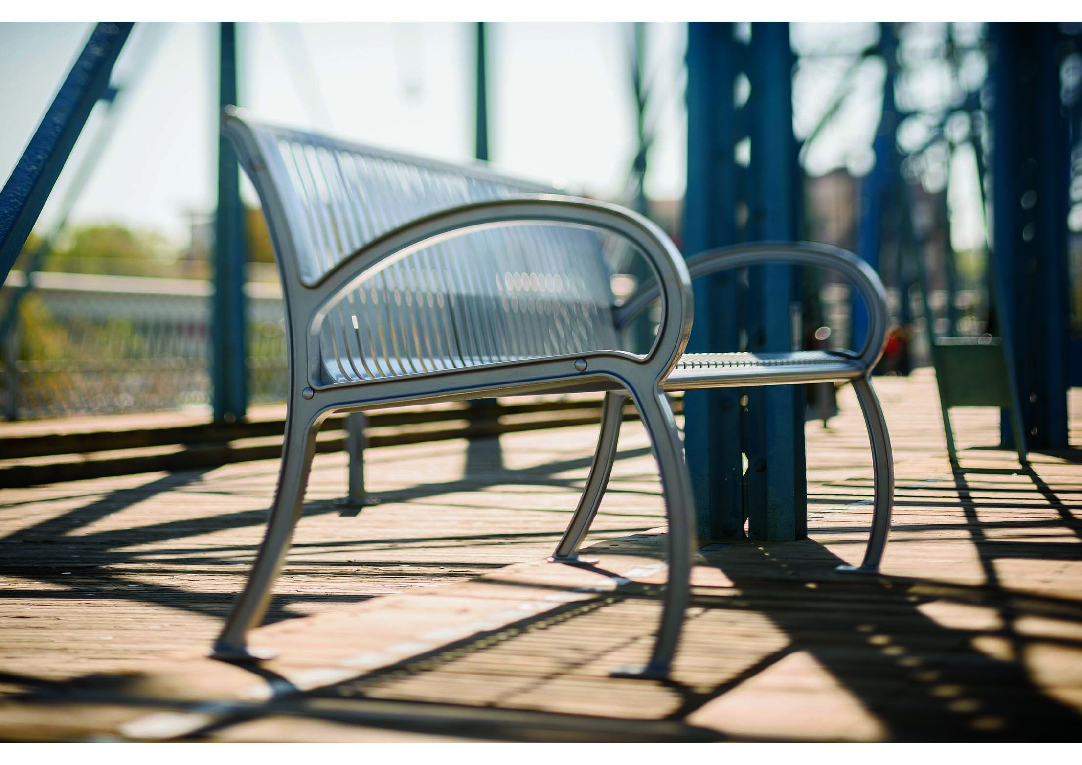 Metal outdoor bench with vertical slats, curved armrests, and cast aluminum frame on wooden deck with blue metal beams