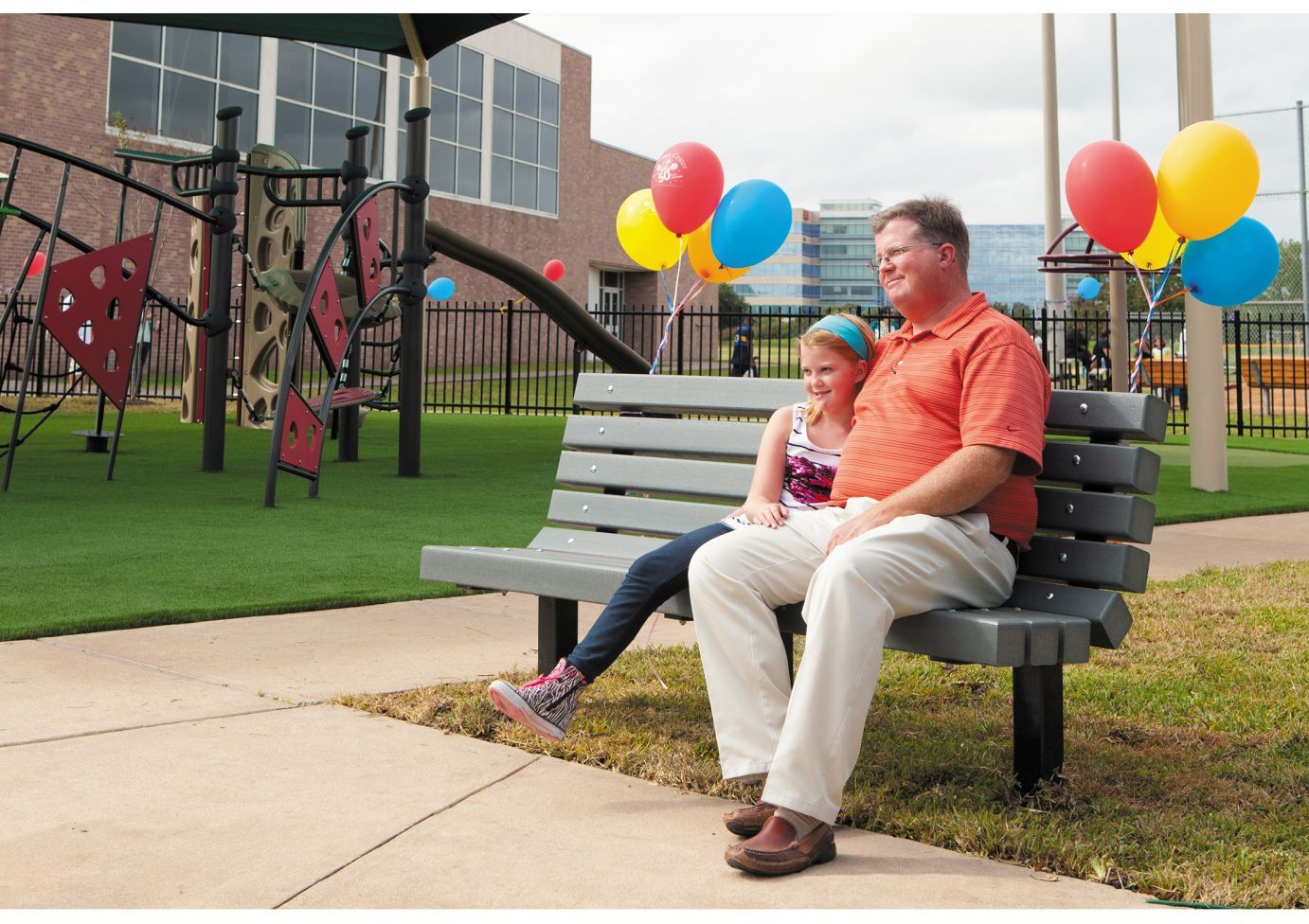 Outdoor recycled plastic bench with backrest occupied by a man and child near playground decorated with colorful balloons