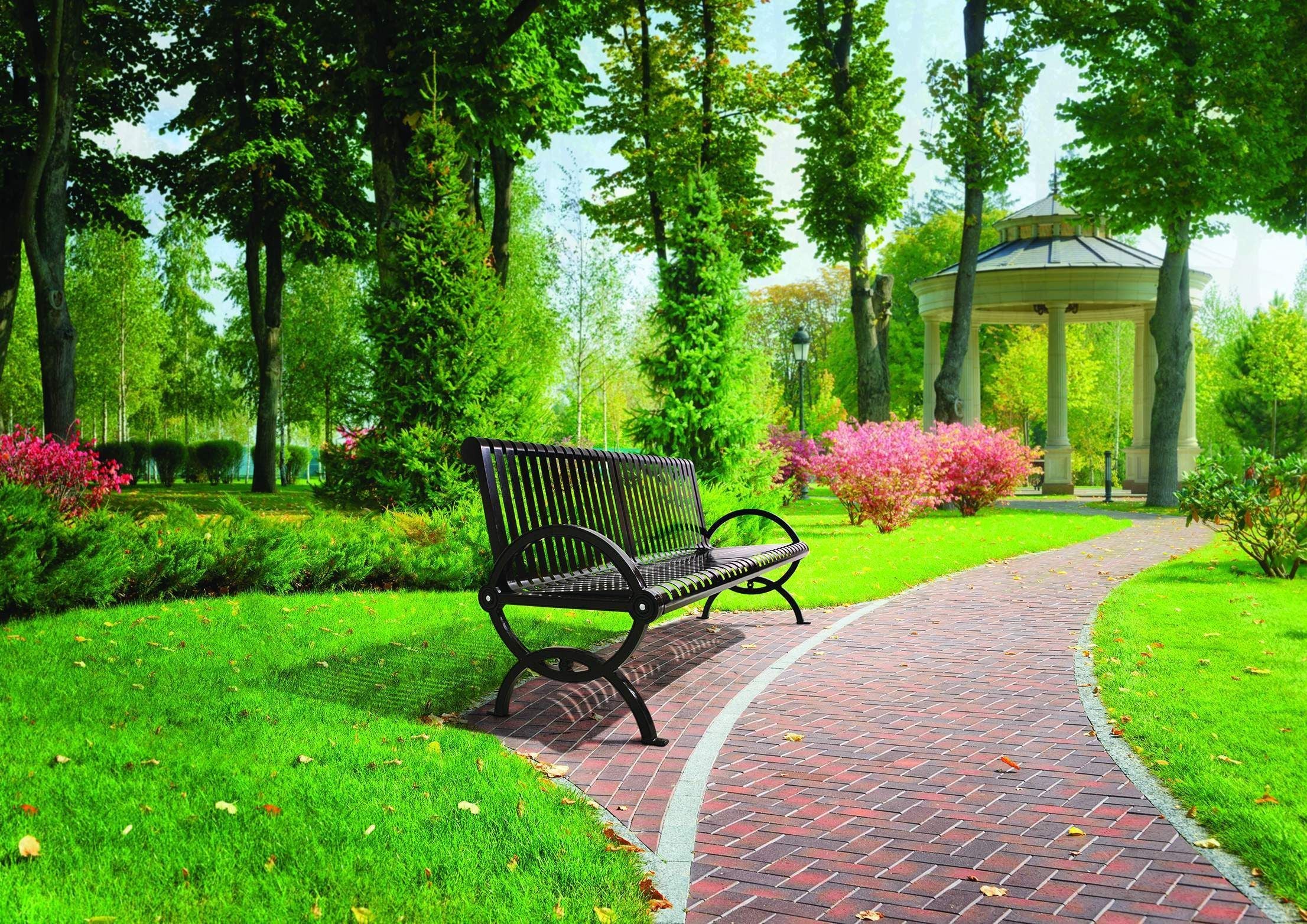 Black metal outdoor bench with vertical slats and curved armrests along a brick pathway in a green park with trees and flowering shrubs