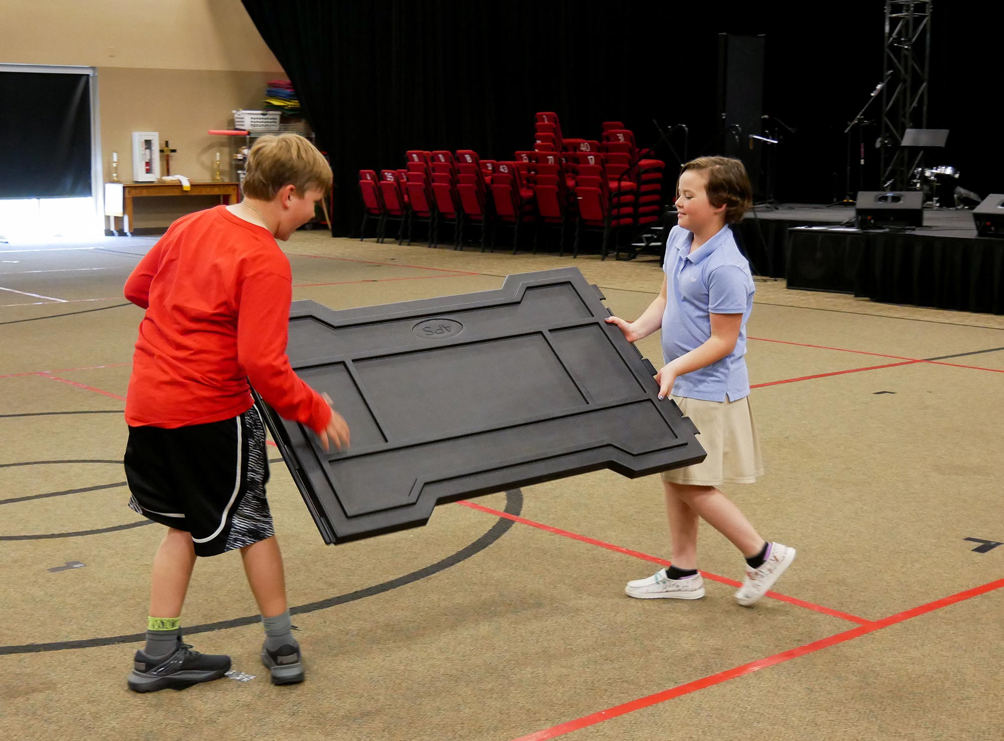 Two children carrying a black foldable portable GaGa Ball pit panel on an indoor gym floor with red court lines