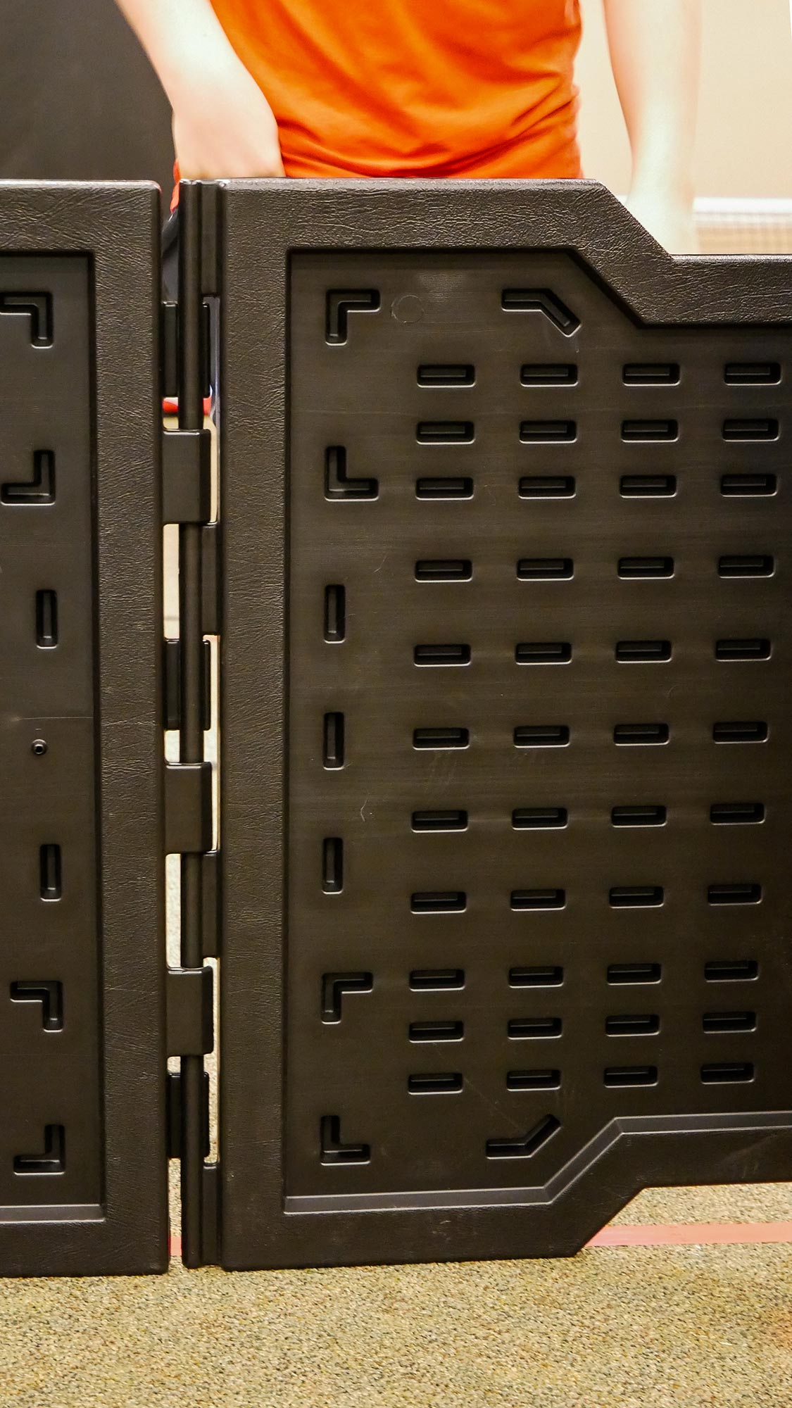 Close-up of black foldable plastic panels with ventilation slots forming a portable GaGa ball pit, person in orange shirt behind