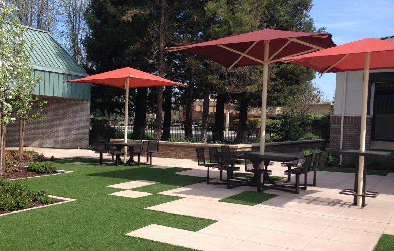 Outdoor patio with red square center post umbrellas shading metal tables and chairs on tiled and artificial grass surfaces