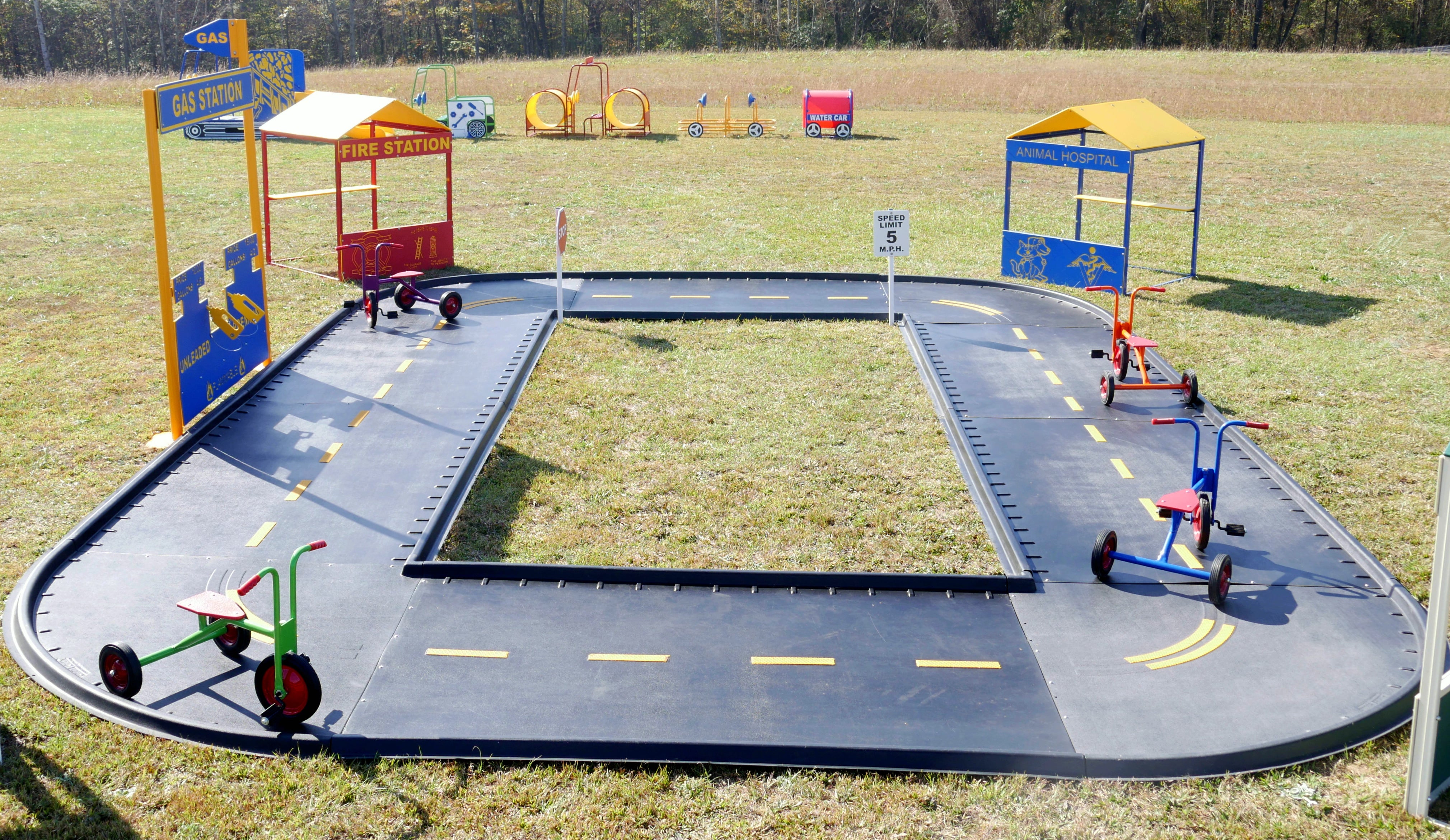 Oval trike path with miniature road signs, labeled shelters, and colorful tricycles on a black track