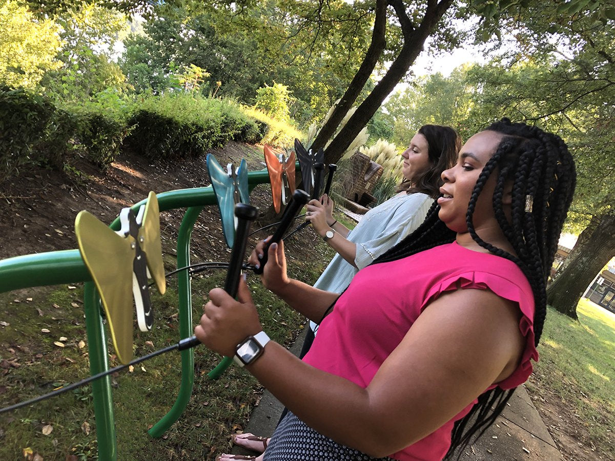 Two women playing colorful butterfly-shaped outdoor musical instruments mounted on a green steel frame in a park setting.
