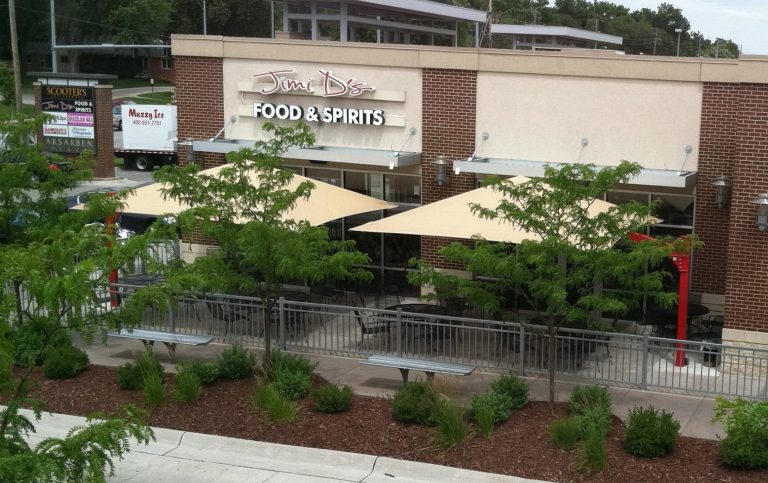 Two beige cantilever square umbrellas shading outdoor dining tables on a restaurant patio with greenery and metal railings