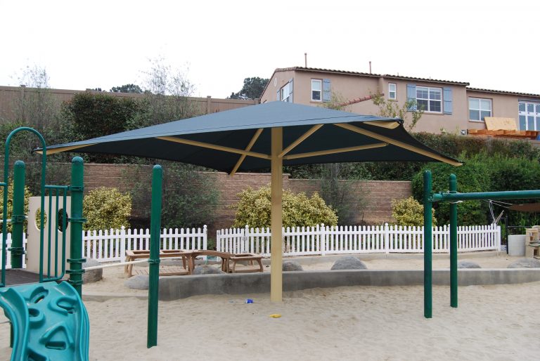 Square center post umbrella with dark fabric and beige frame shading sandy playground area with picnic tables and green play structures