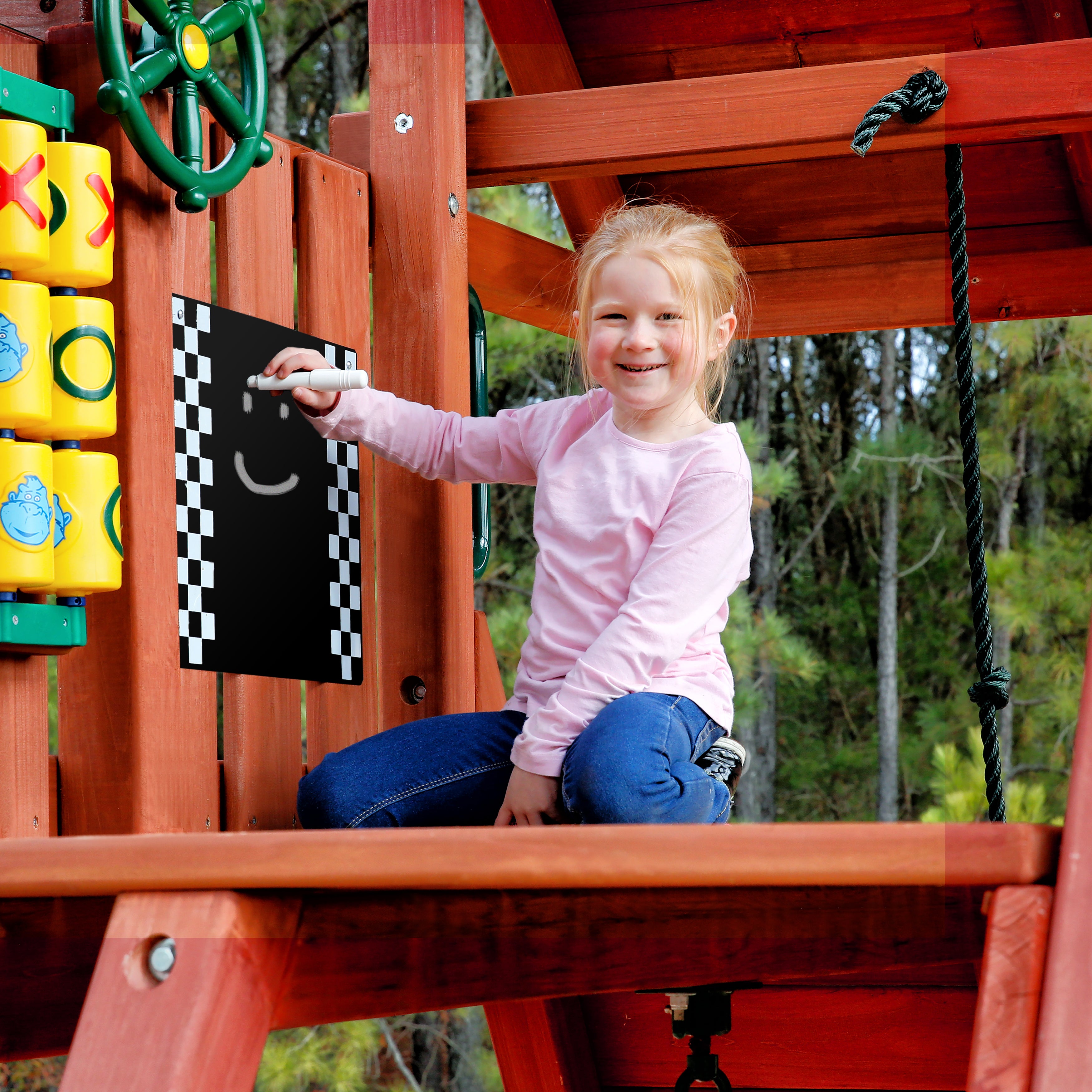 Child drawing a smiley face with chalk on a black metal outdoor chalkboard with checkered border attached to a wooden swing set