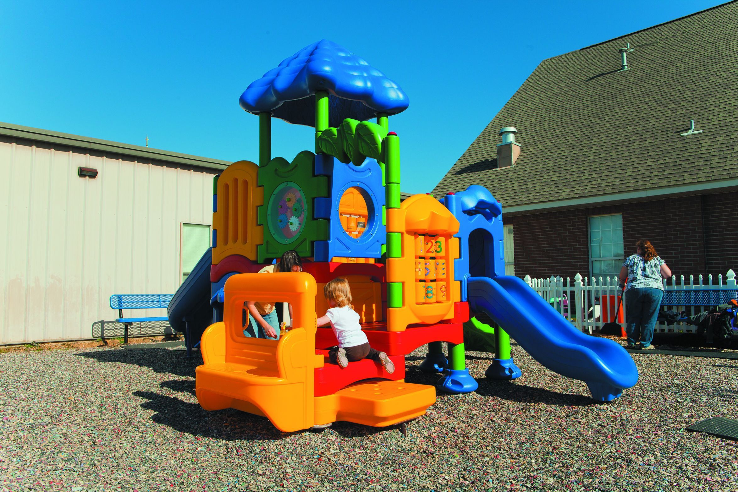 Colorful outdoor playground with roof, slides, interactive panels, and children playing on gravel surface