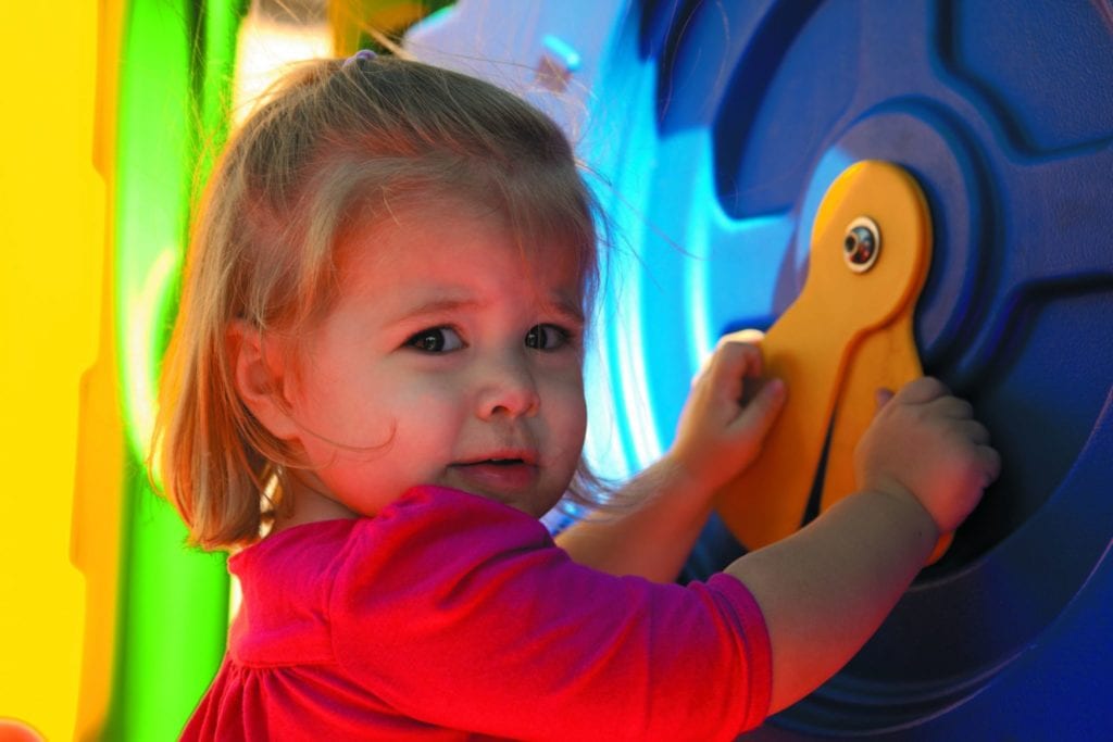 Toddler engaging with colorful playground clock panel featuring large yellow hands on blue background under leafy canopy shade