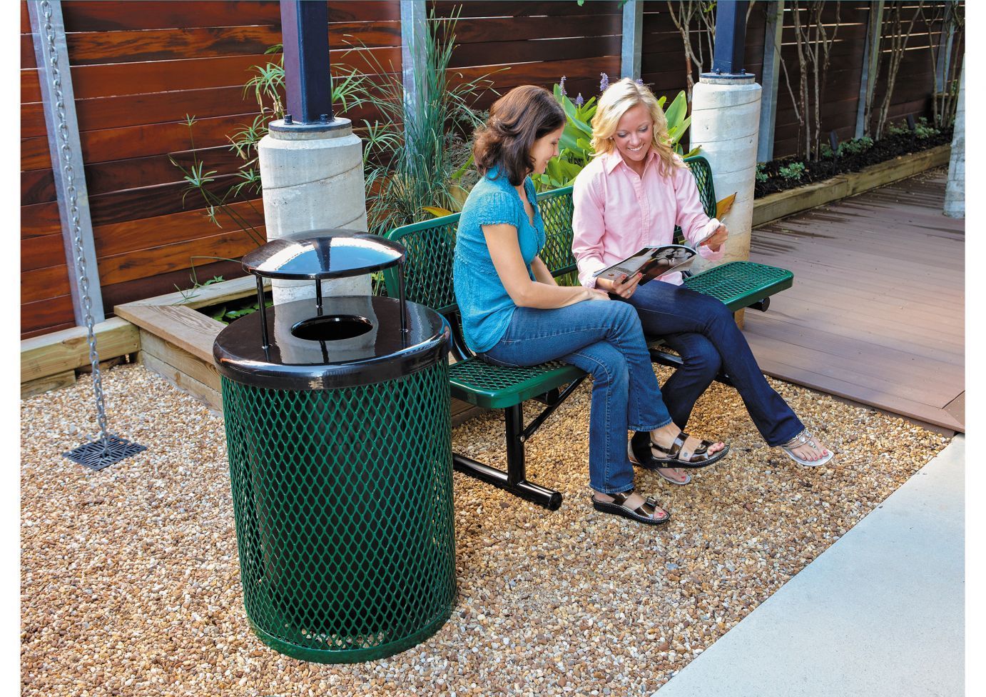 Outdoor green metal trash receptacle with diamond pattern and round dome lid near two women sitting on matching green metal bench