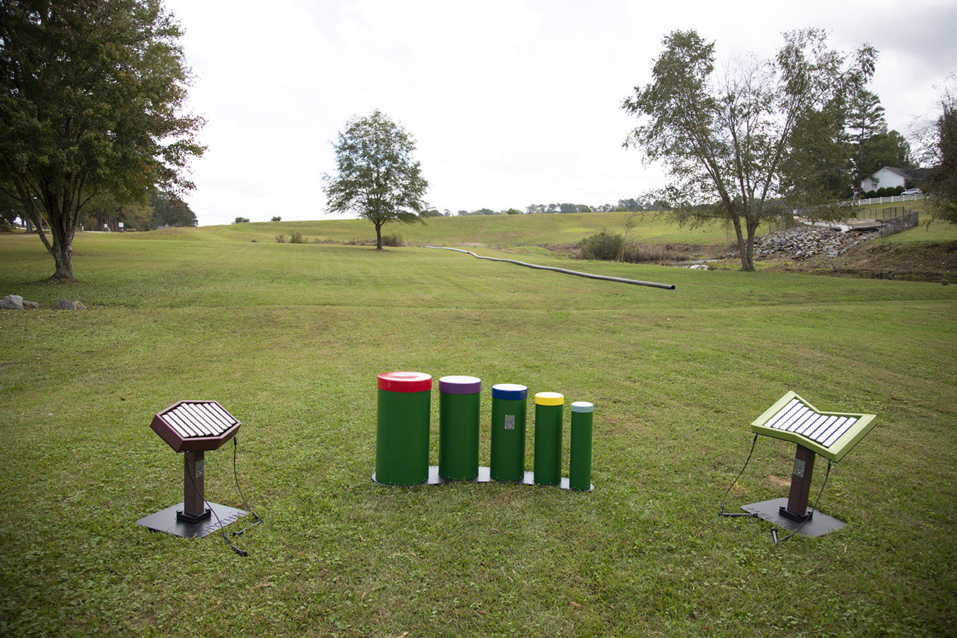 Toddler-height outdoor musical instruments including tuned drums and xylophones on grass in a park setting