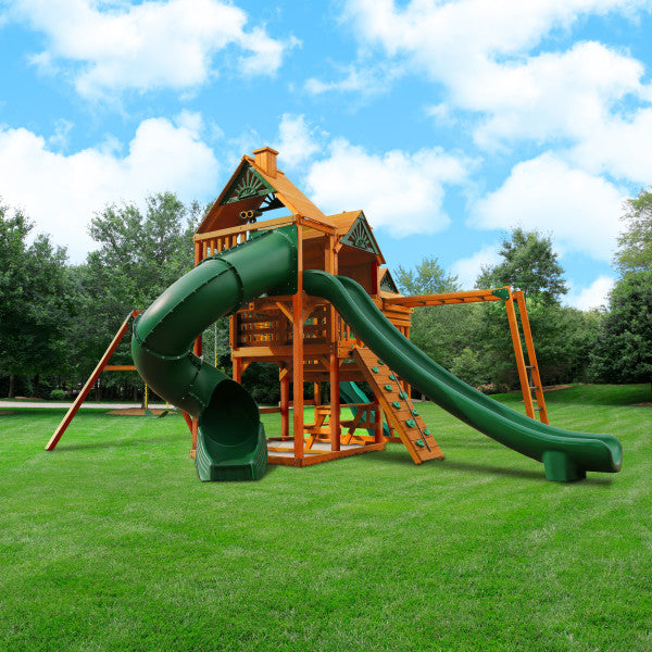 Cedar wooden playset with three decks, green tube and wave slides, rock climbing wall, monkey bars, and swings on grass lawn