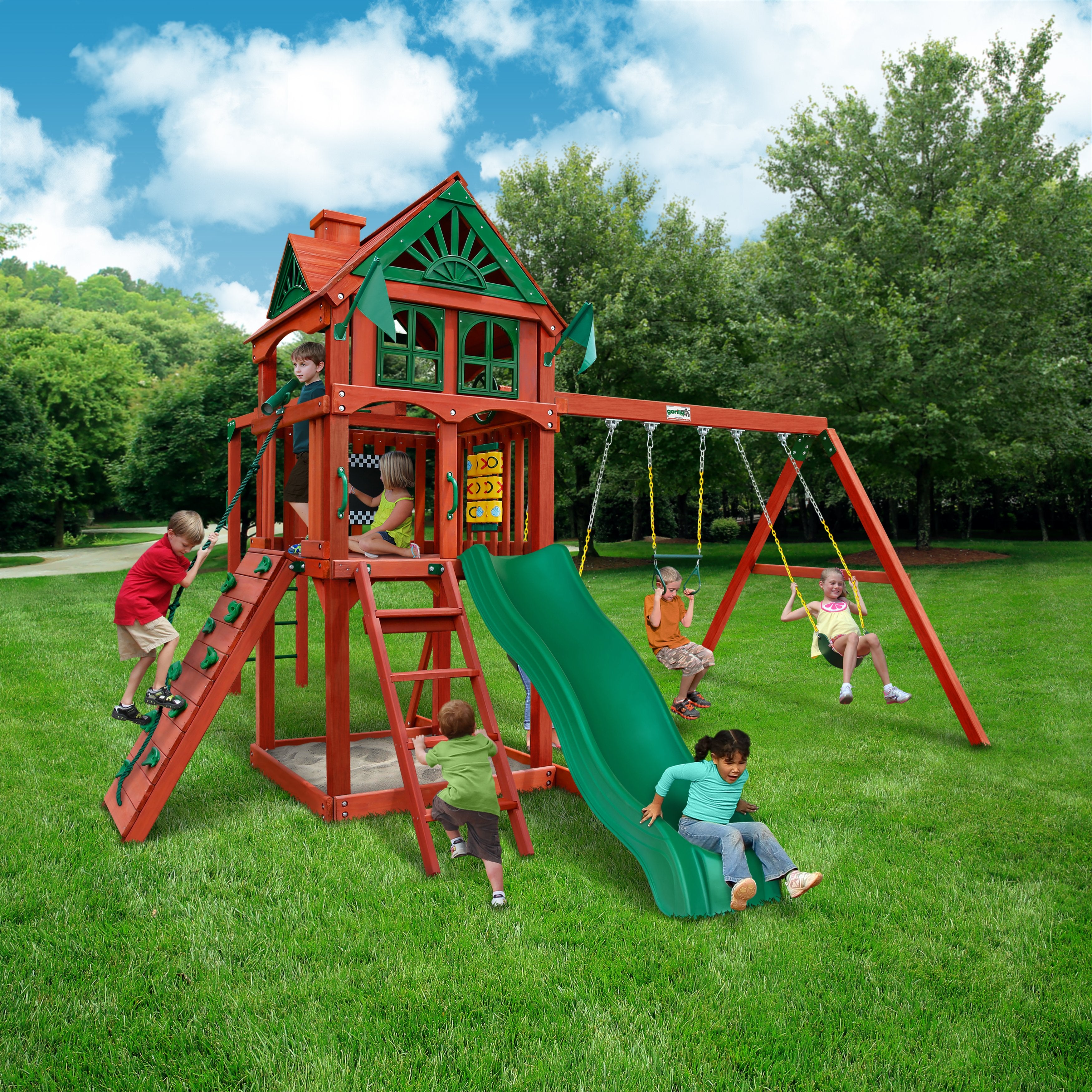 Cedar wooden playset with green wave slide, swings, rock climbing wall, sandbox, and clubhouse with decorative windows, children playing outdoors
