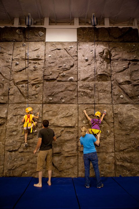 Indoor modular climbing wall with colorful handholds and textured panels, children climbing under adult supervision