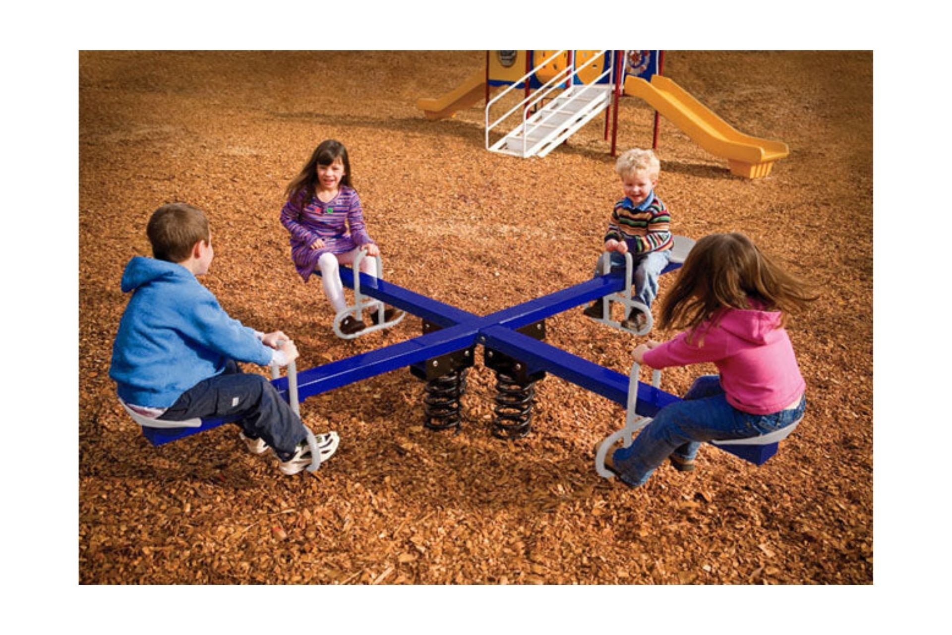 Four children seated on a blue four-seat spring rider with white handles on a wood chip playground surface
