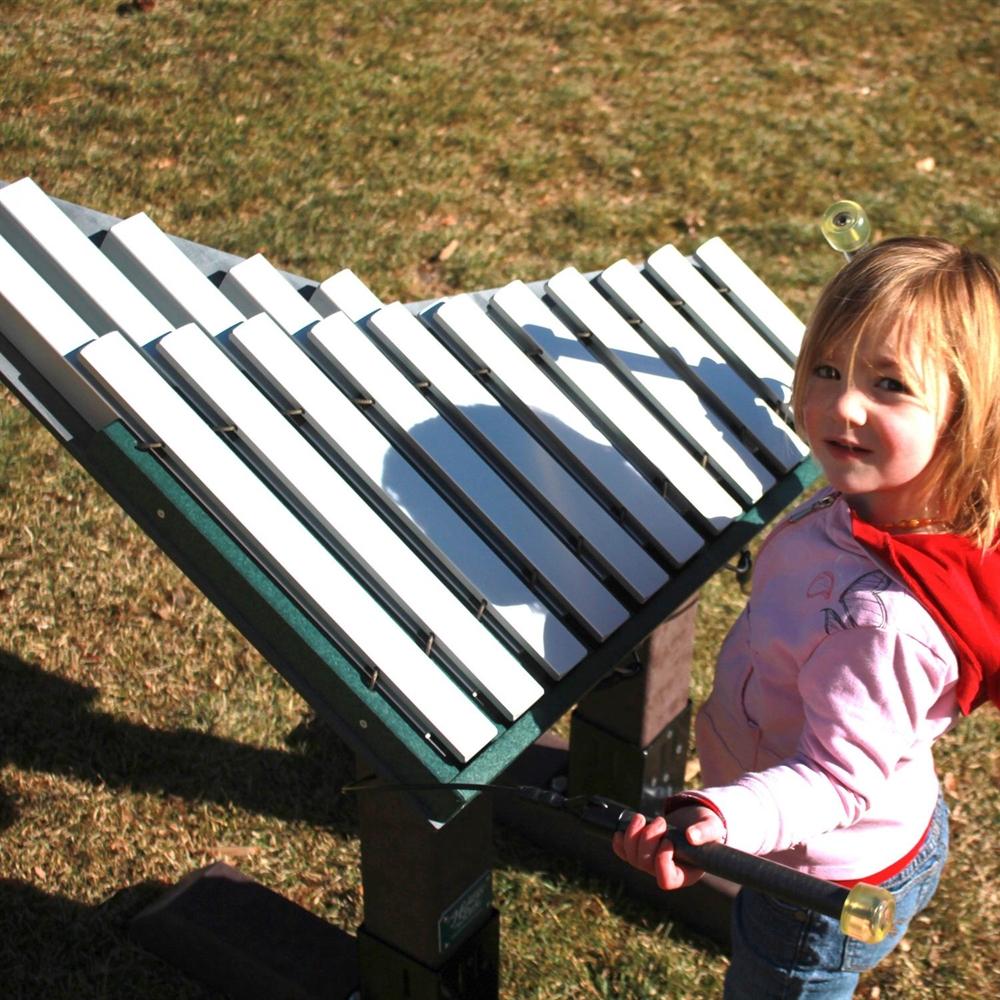 Green outdoor metallophone with white bars, mallets in embedded slots, child holding mallets on grass