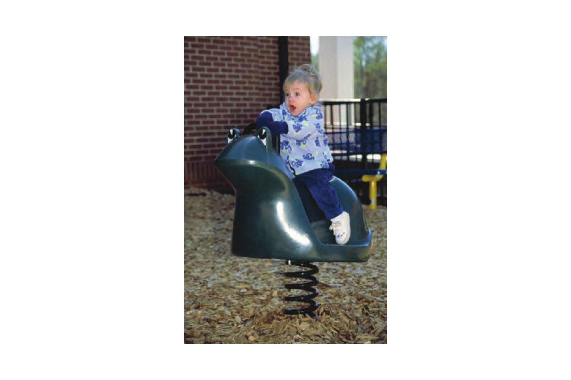 Frog Molded Spring Rider on wood chip playground surface with child playing near brick wall and bench