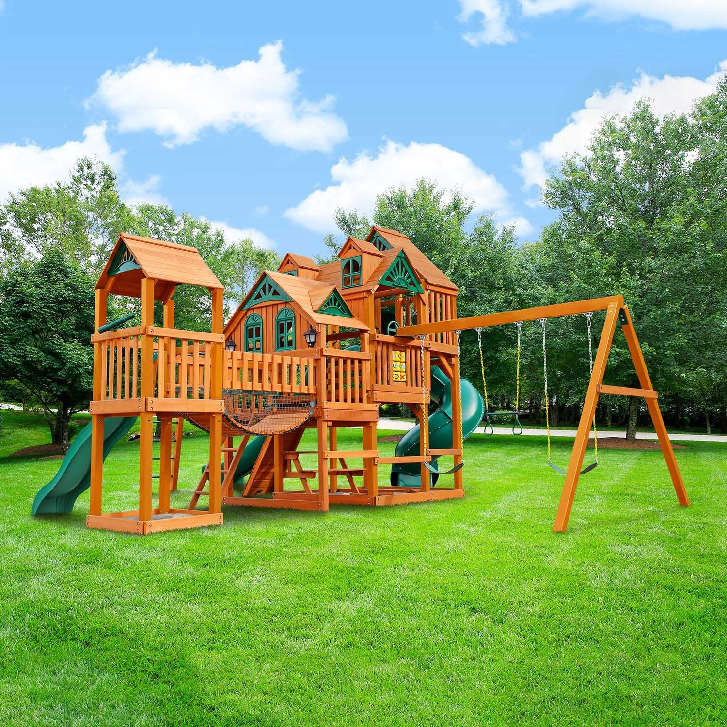 Wooden outdoor playset with two forts, green slides, climbing wall, swings, monkey bars, tic-tac-toe panel, and picnic table on grass