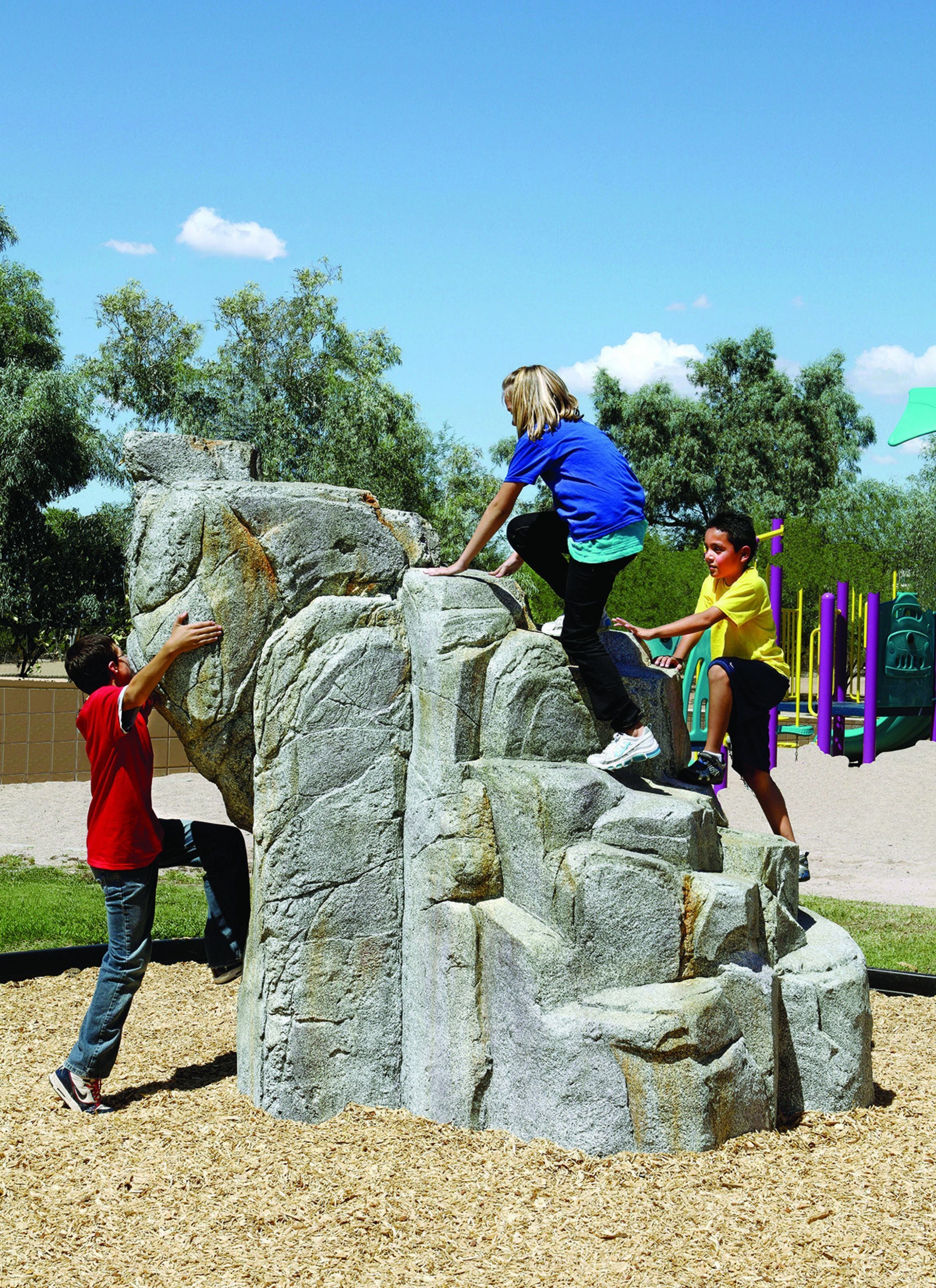 Textured granite climbing boulder with three children climbing, surrounded by playground mulch and equipment