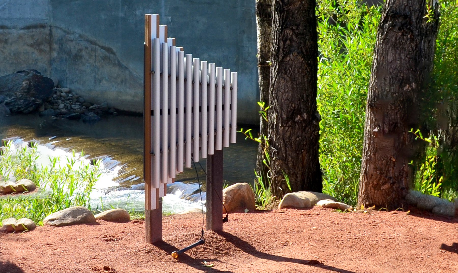 Griffin Outdoor Musical Park Instrument with anodized aluminum chimes, tan frame, set outdoors near trees and water