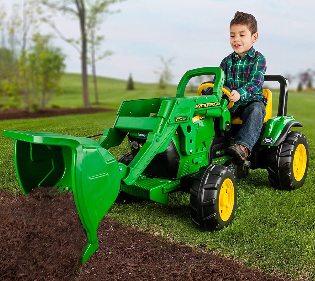 Child on green and yellow pedal tractor with large front loader scooping mulch on grass outdoors