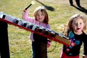 Outdoor 18-note metallophone with blue trim and mallet holders, played by children in grassy park setting