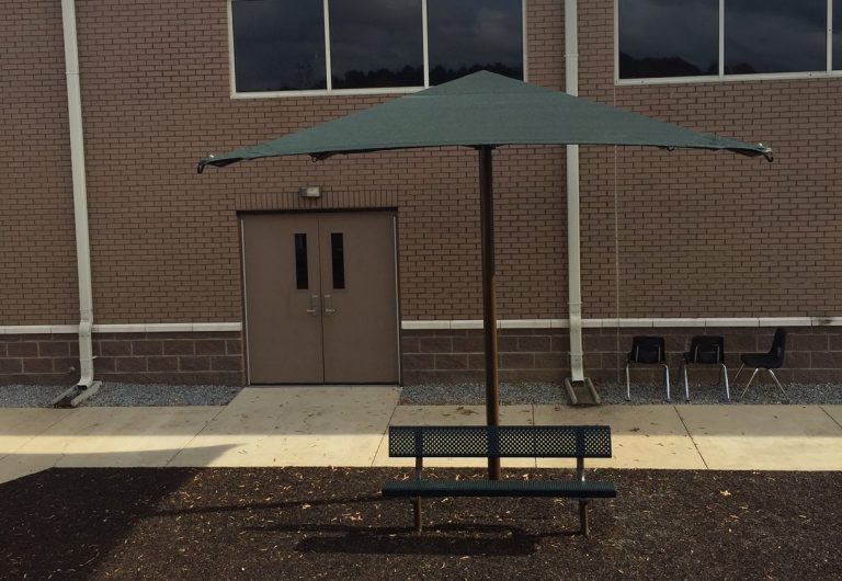 Square green fabric center post umbrella shade over metal bench in outdoor playground setting