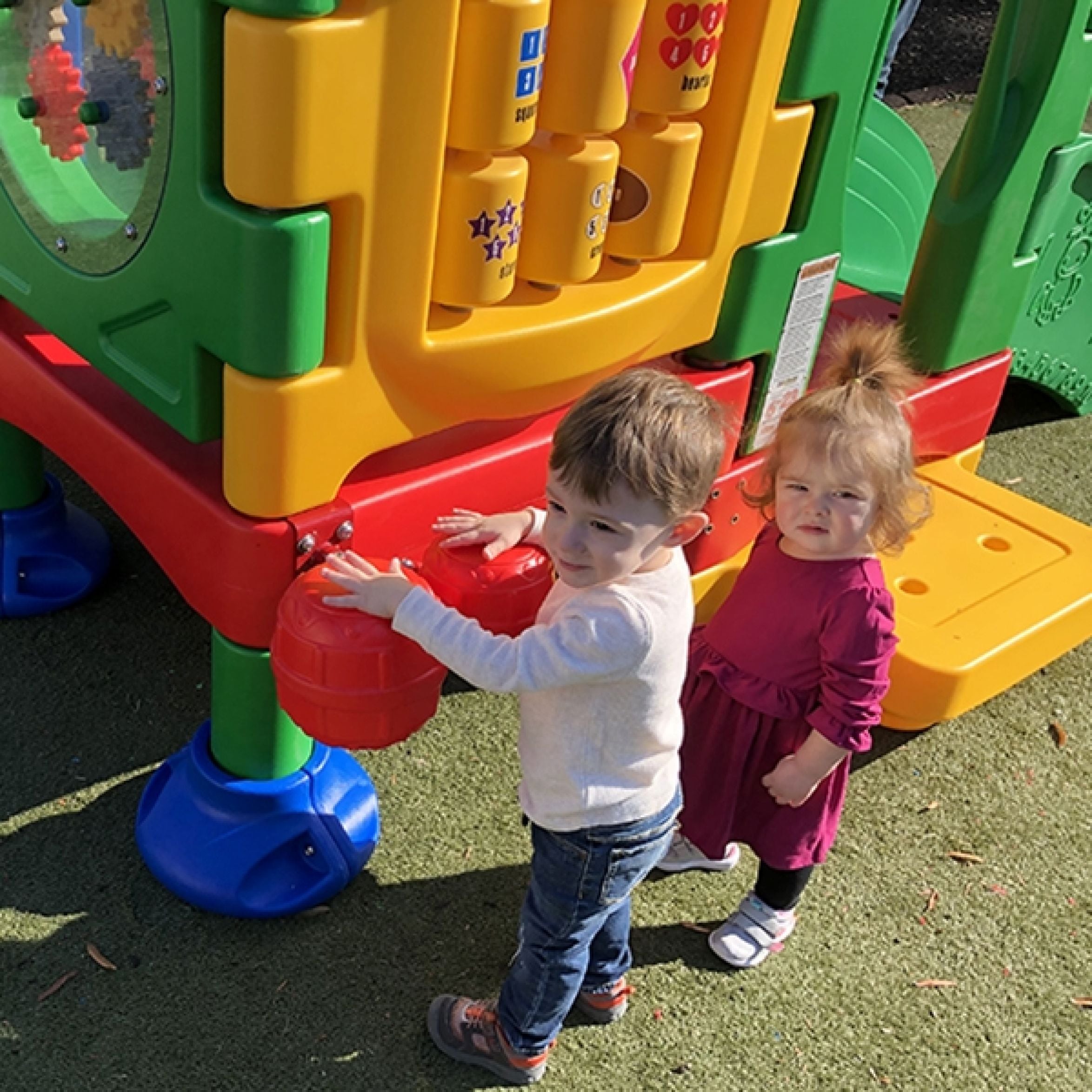 Two toddlers playing on a colorful two-deck plastic playground with interactive panels, musical bongo drums, and animal images