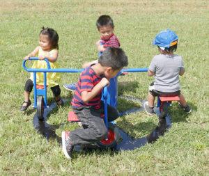 Colorful four-seat inground play rider with children pedaling together in a circular motion on grass