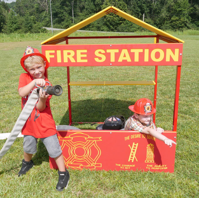 Two children wearing firefighter helmets playing with a colorful fire station playhouse on grass, one holding a hose.