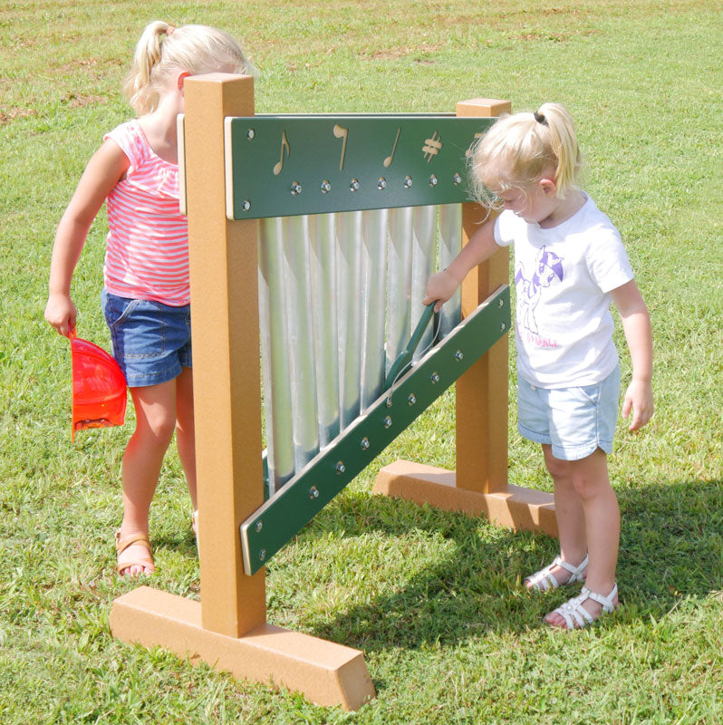 Two young girls playing a colorful freestanding outdoor chime wall with metal tubes and musical note decorations on grass