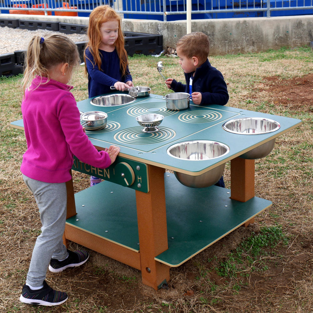 Outdoor children's mud kitchen with green tabletop, stainless steel bowls and pots, and three kids playing around it