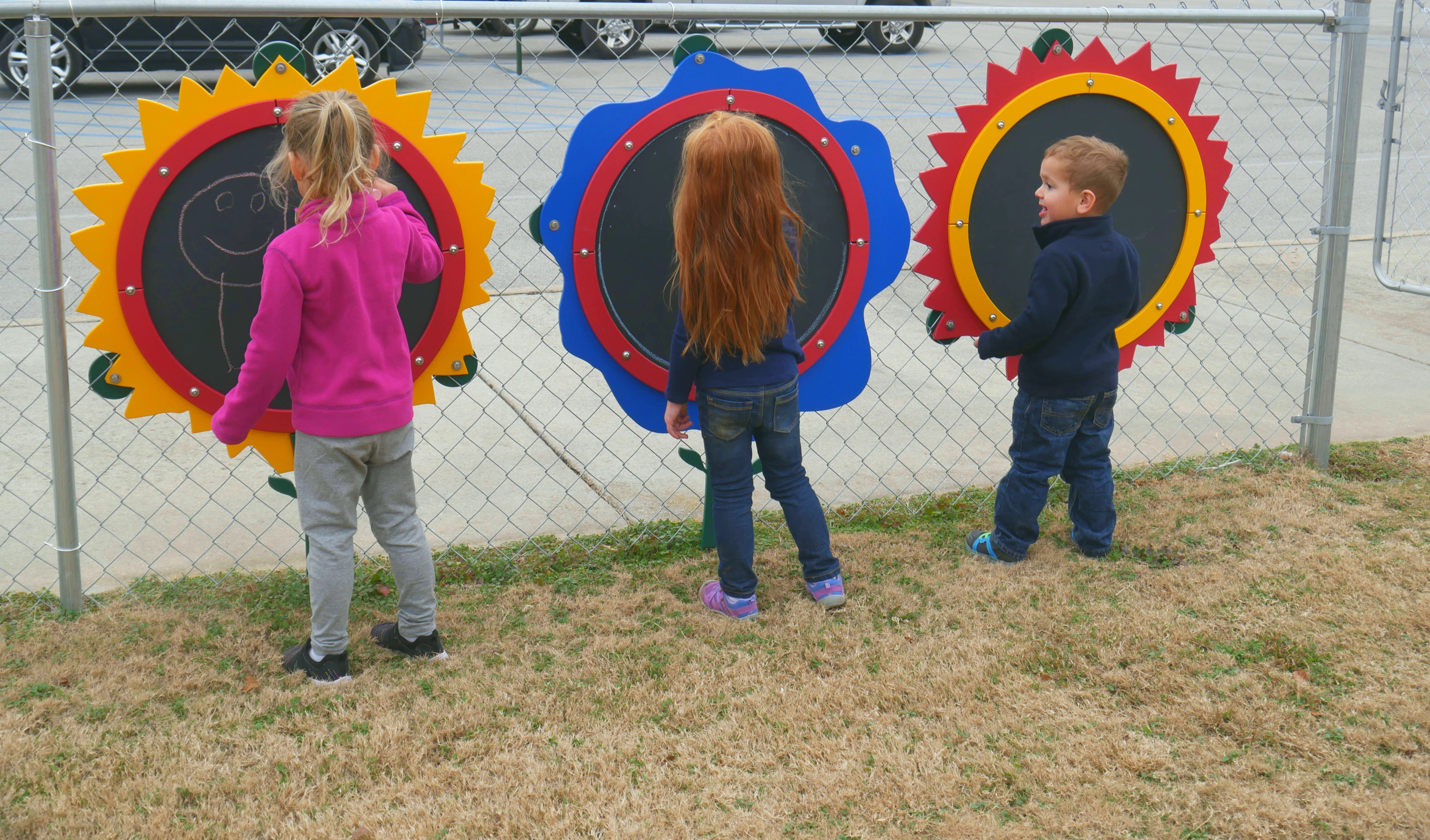Three children engaging with colorful flower-shaped outdoor chalkboards mounted on a chain-link fence, promoting creative play and learning.