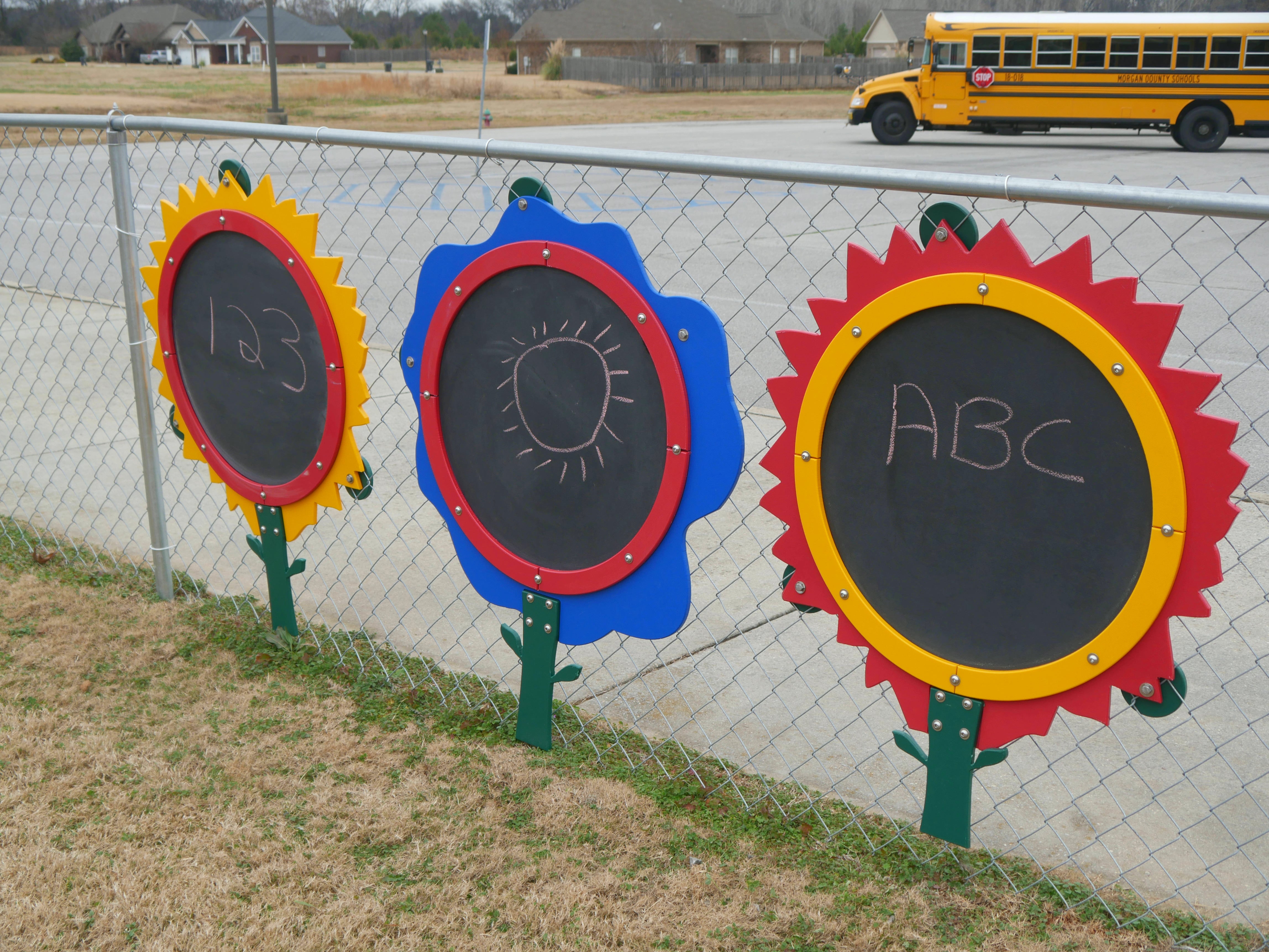 Three colorful flower-shaped outdoor chalkboards with chalk drawings mounted on a chain-link fence
