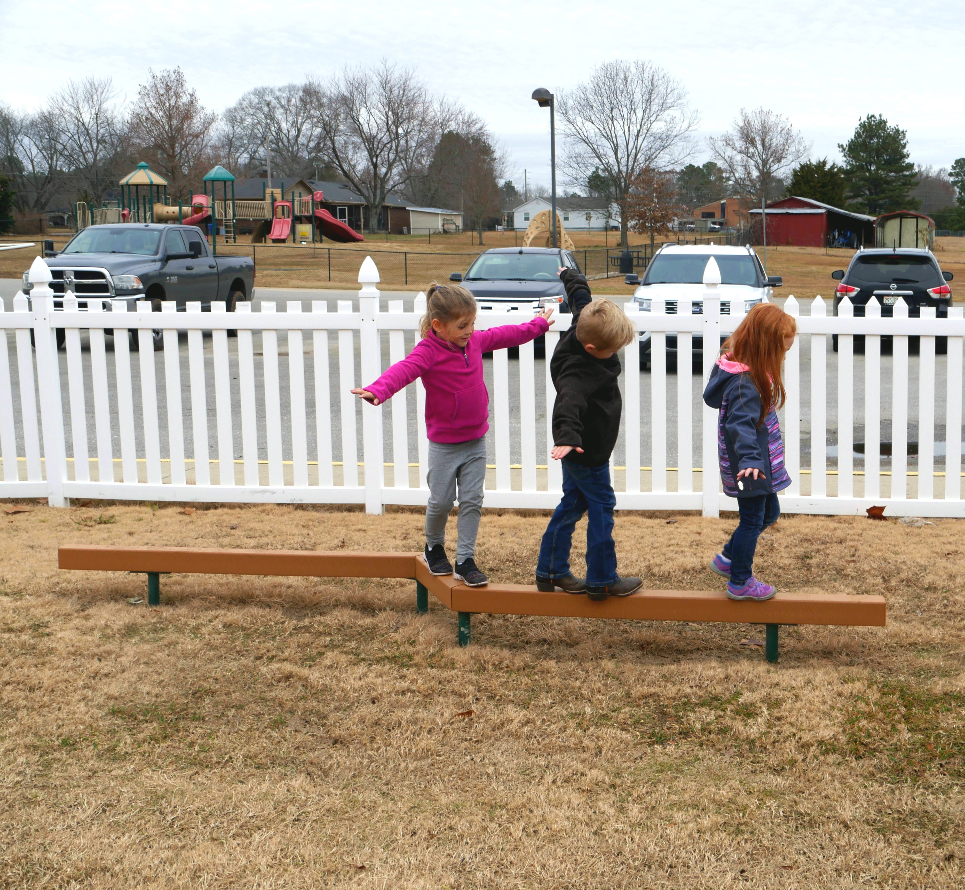 Three children balancing on a brown recycled plastic lumber zig zag balance beam outdoors on grass
