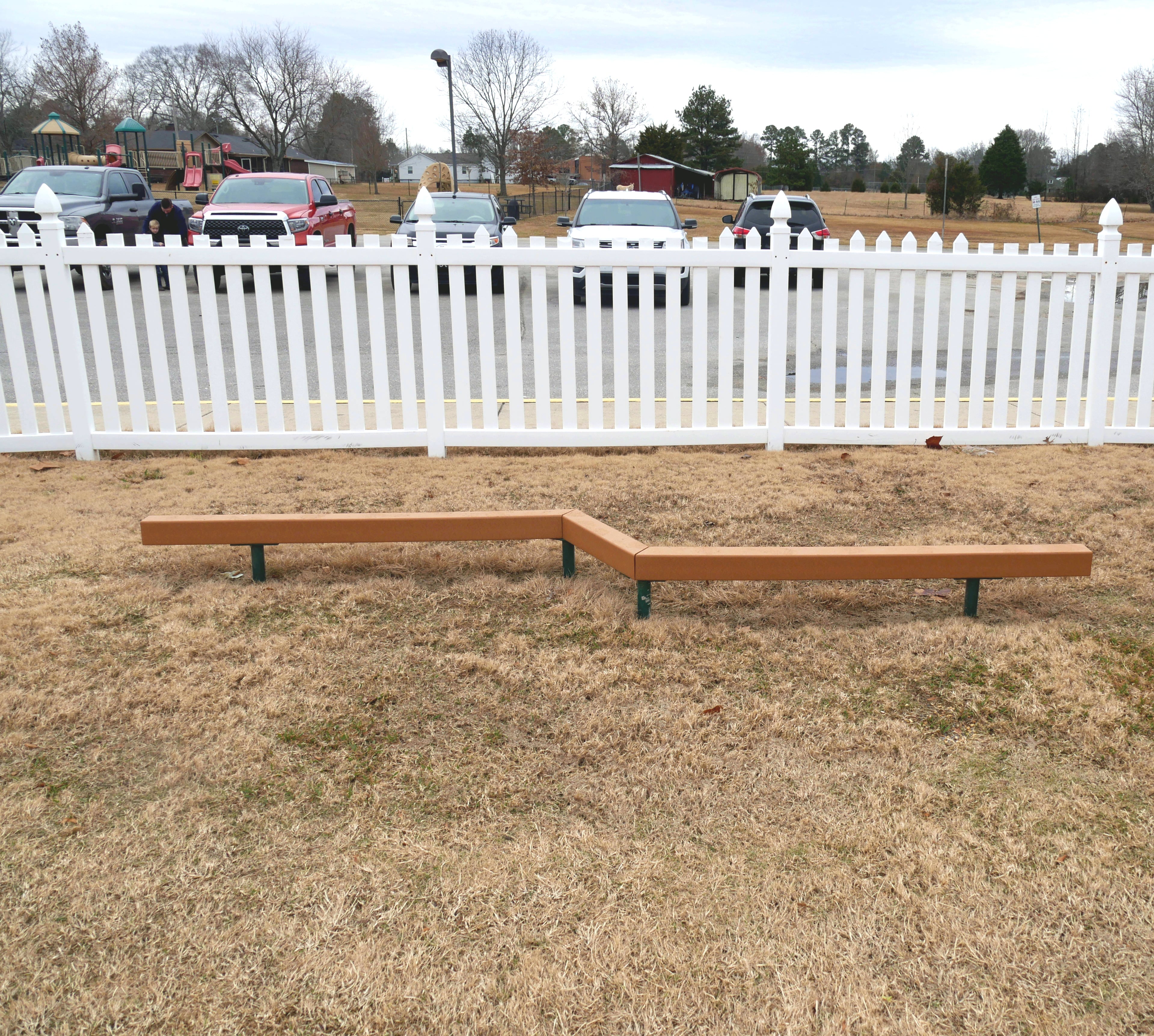 Outdoor zigzag balance beam made of brown recycled plastic lumber on dry grass near a white picket fence