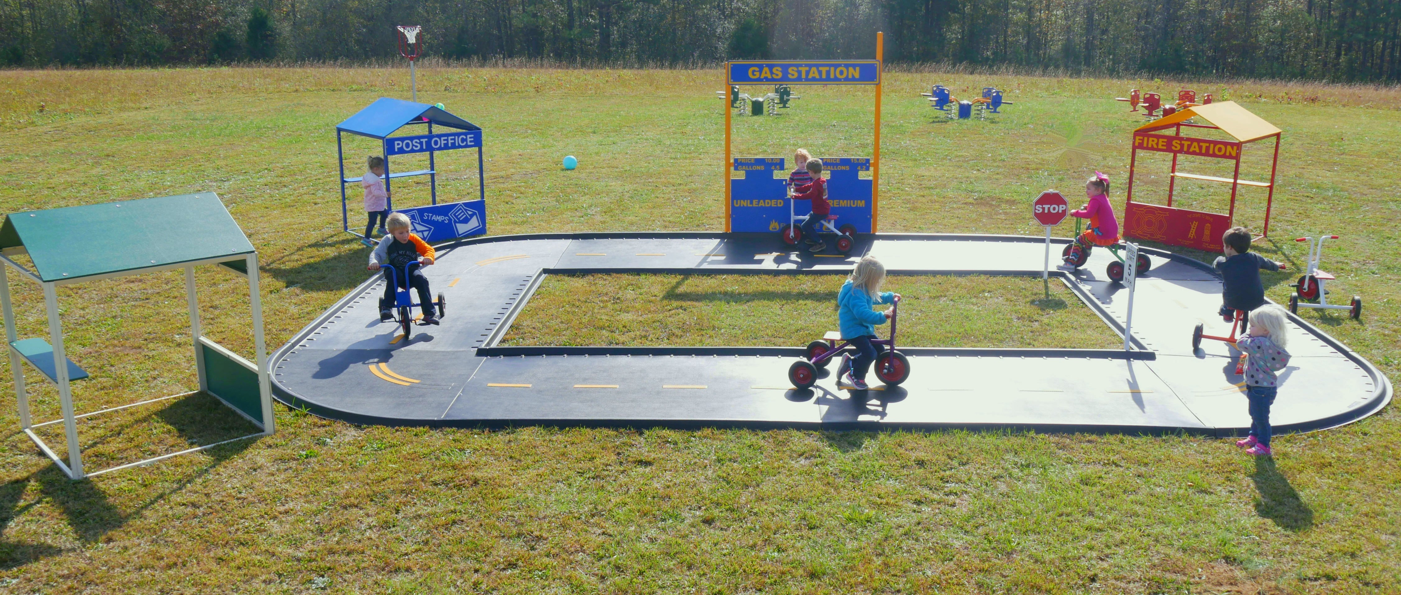 Oval trike path with road markings, miniature gas station, post office, fire station, and children riding tricycles outdoors