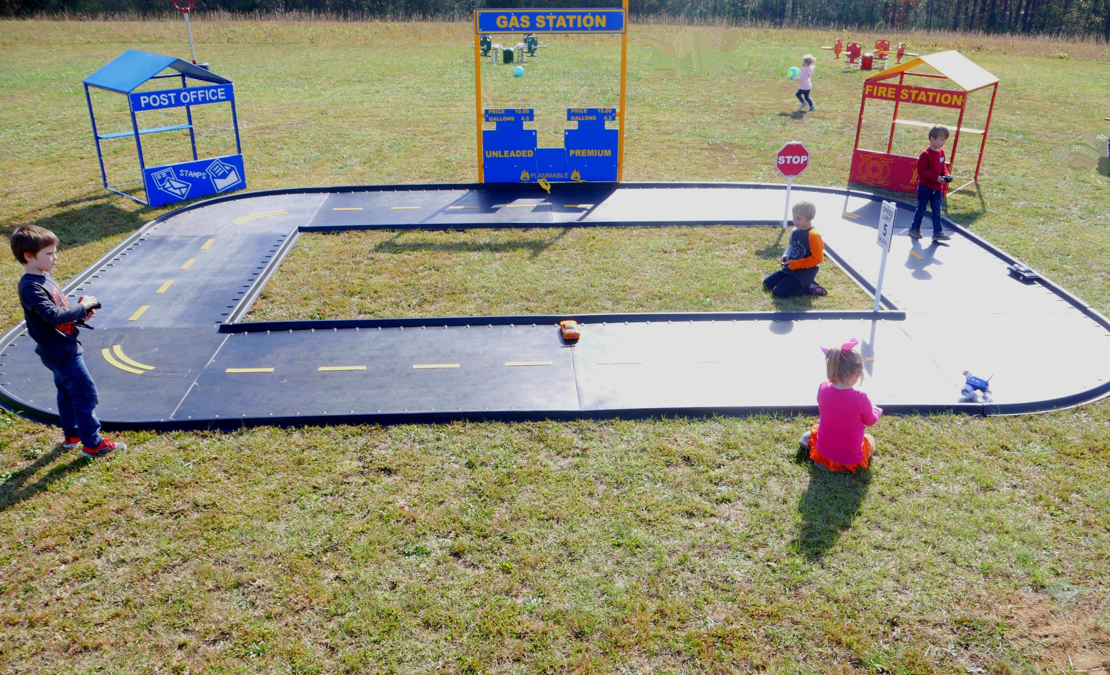 Oval trike path with miniature road signs, play buildings, and children using remote control cars on grass