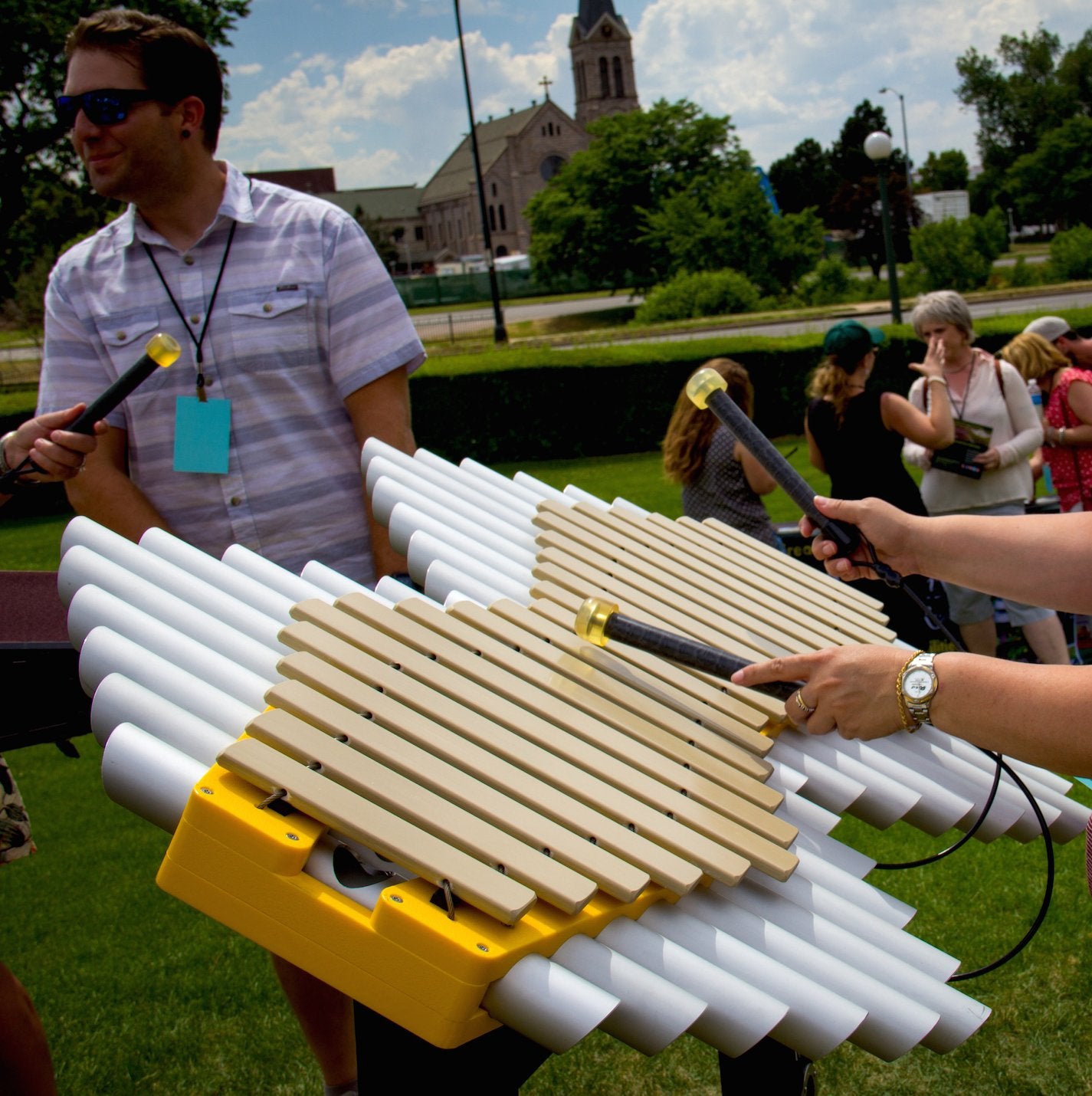 Outdoor yellow pentatonic Imbarimba with beige fiberglass bars and white resonator tubes played by two people using mallets
