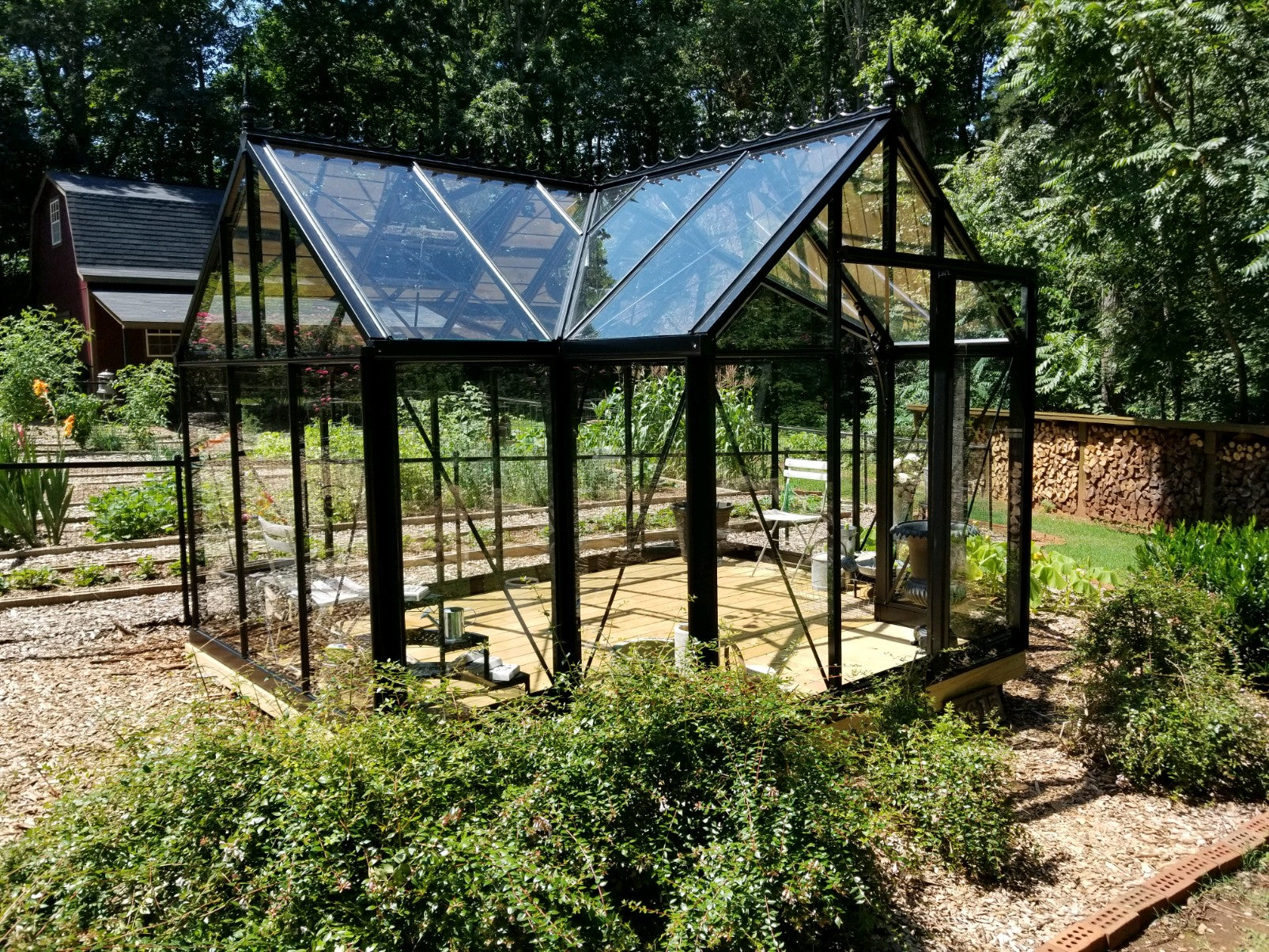 Black-framed glass greenhouse with pitched roof, automatic vents, sliding door, surrounded by garden beds and greenery