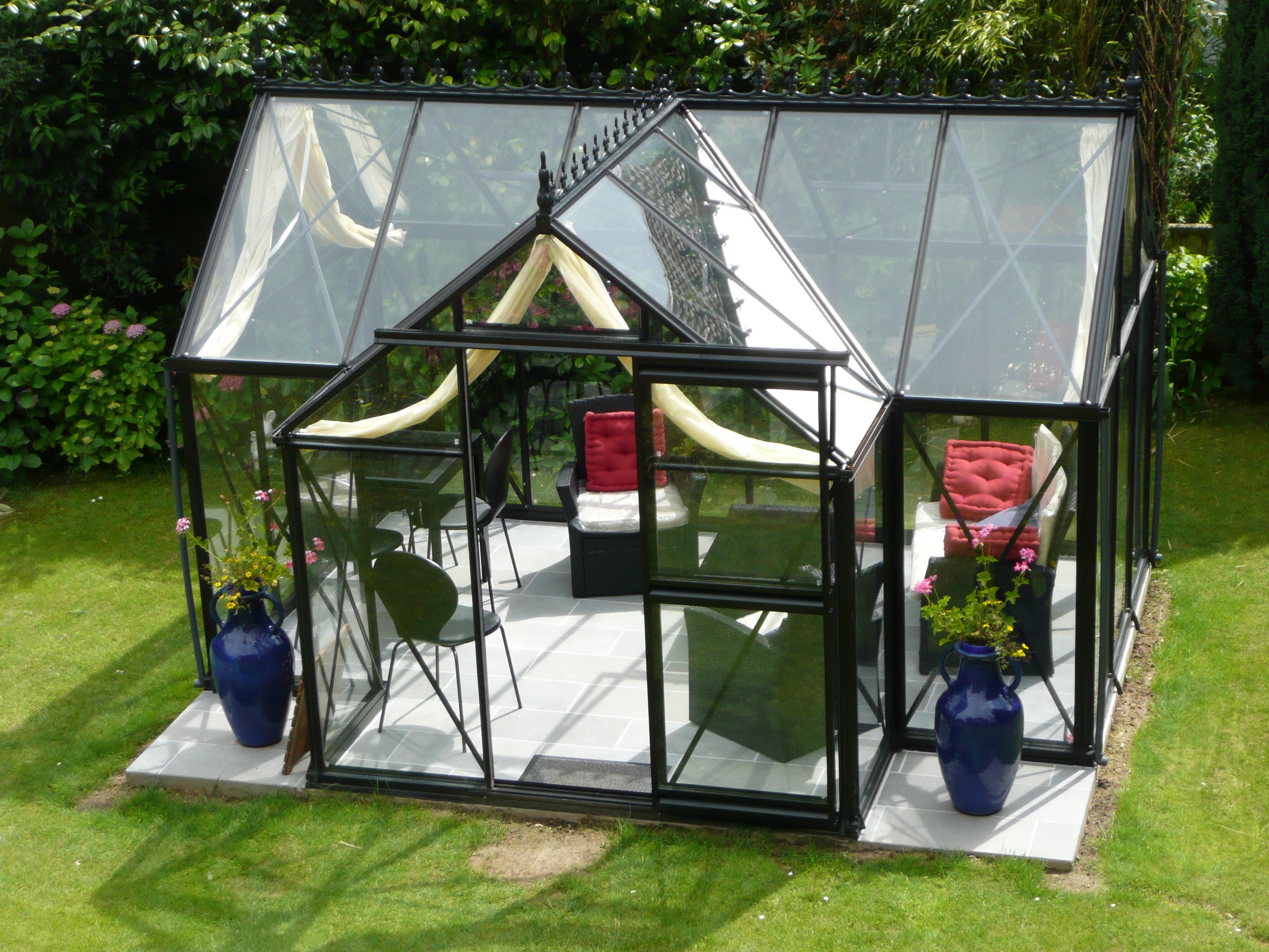 Junior Orangerie Greenhouse with elegant black frame, glass panels, roof windows, sliding door, and garden seating with red cushions