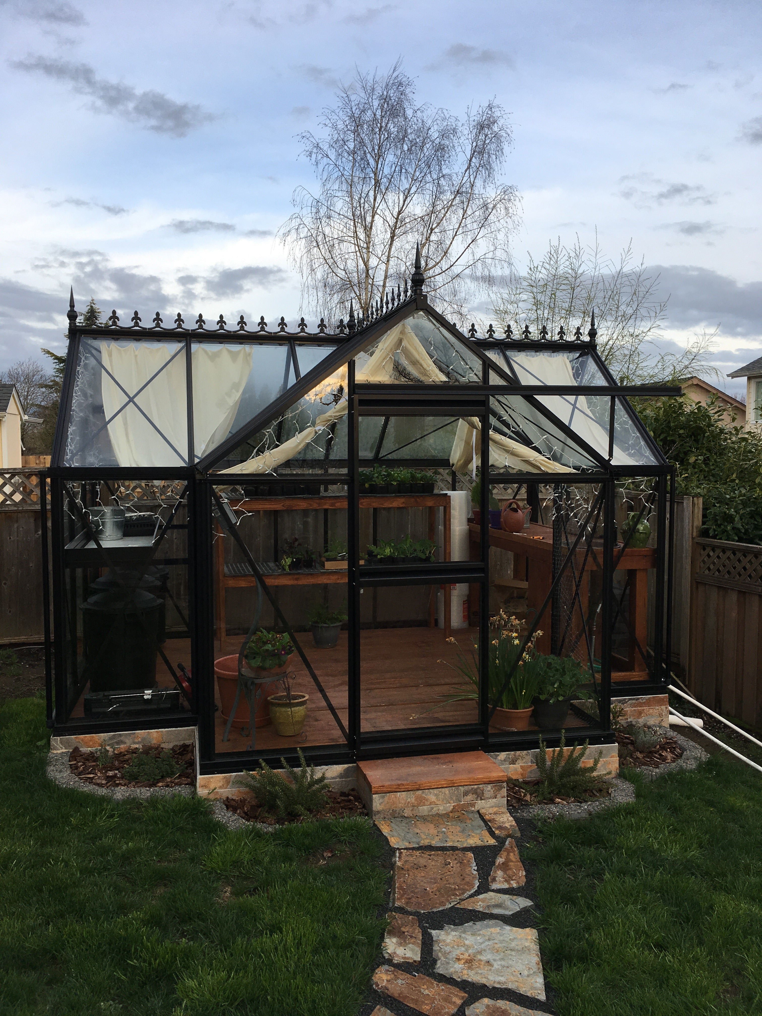Black-framed Junior Orangerie greenhouse with decorative roof finials, glass panels, interior plants, wooden floor, and stone path entrance
