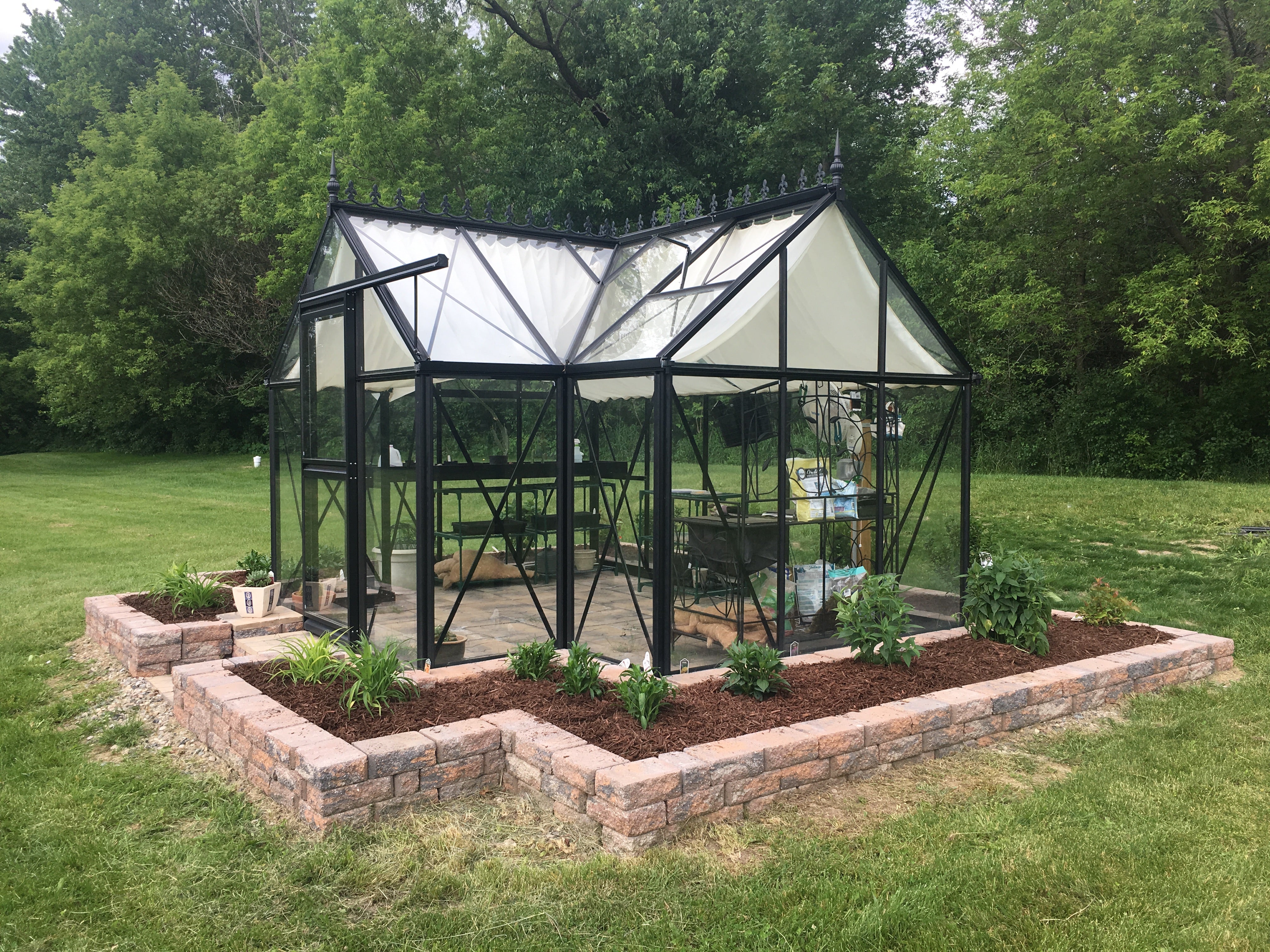 Black-framed peaked-roof greenhouse with sliding door, roof vents, and surrounding raised garden beds with plants