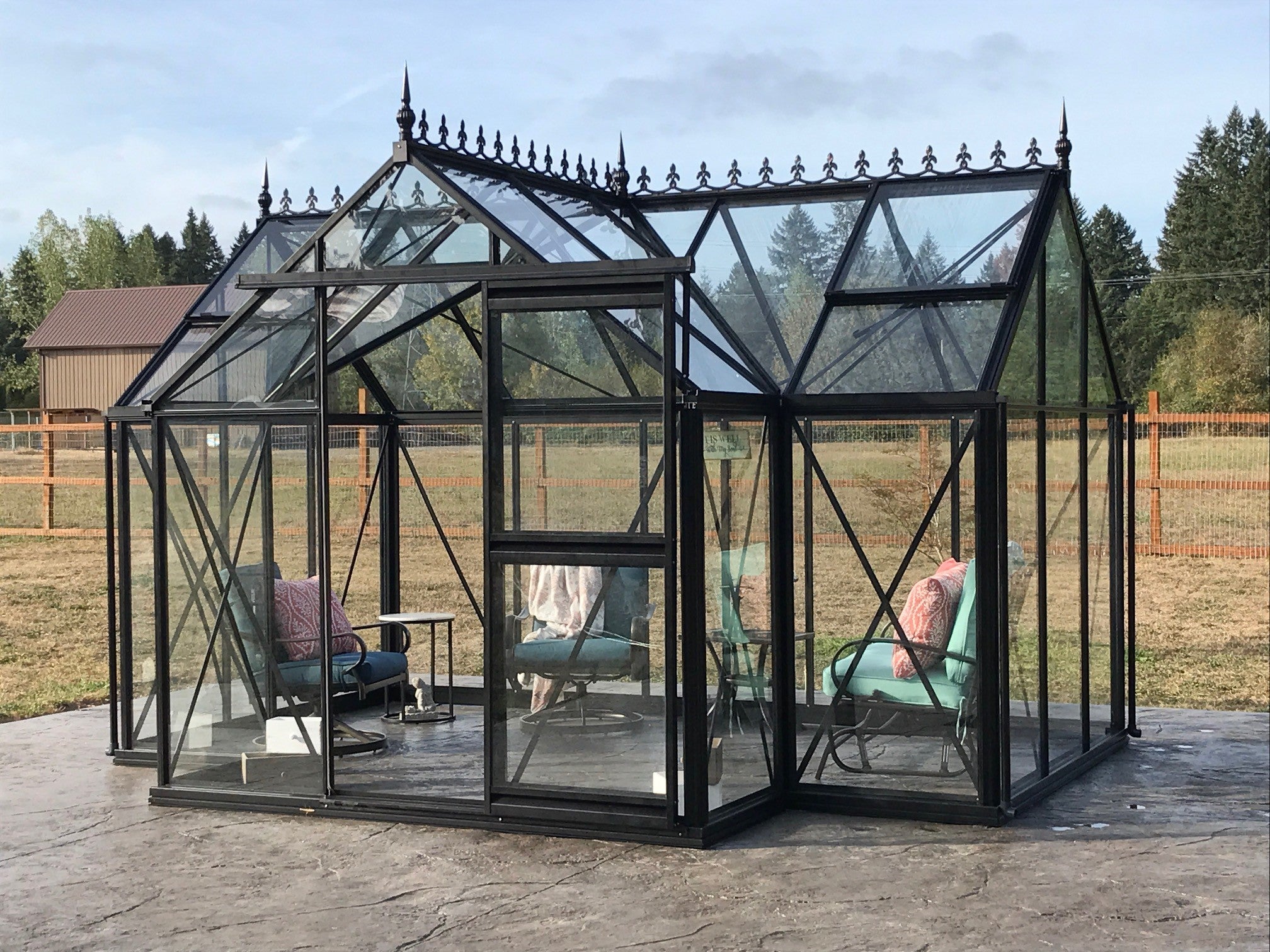 Black-framed glass greenhouse with ornate roof finials, two roof vents, sliding glass door, and cushioned chairs on concrete base