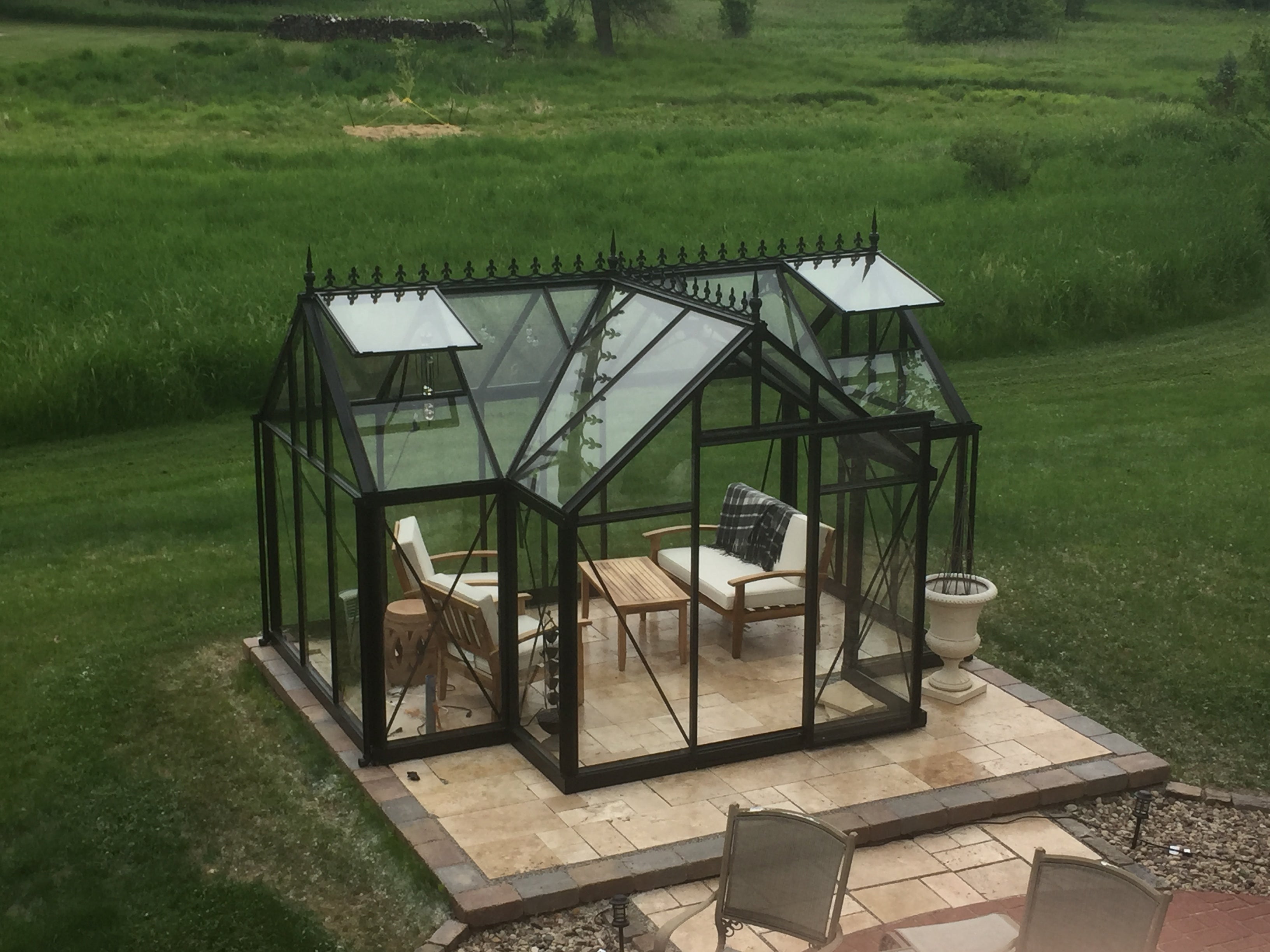 Black-framed Junior Orangerie greenhouse with decorative roof finials, two open roof windows, sliding glass door, and patio furniture on stone tiles
