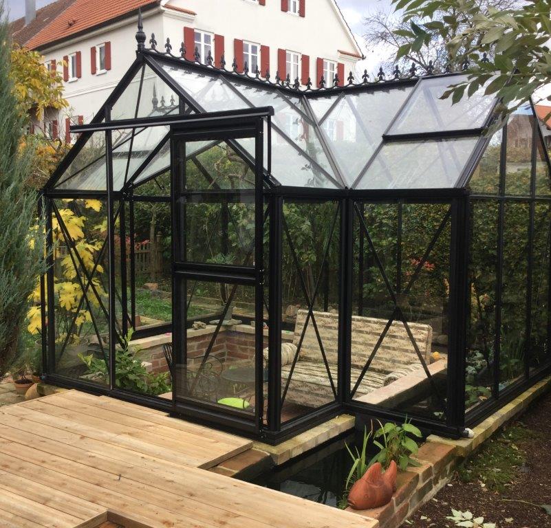 Black-framed Junior Orangerie greenhouse with glass panels, decorative roof finials, sliding door, and interior seating surrounded by plants