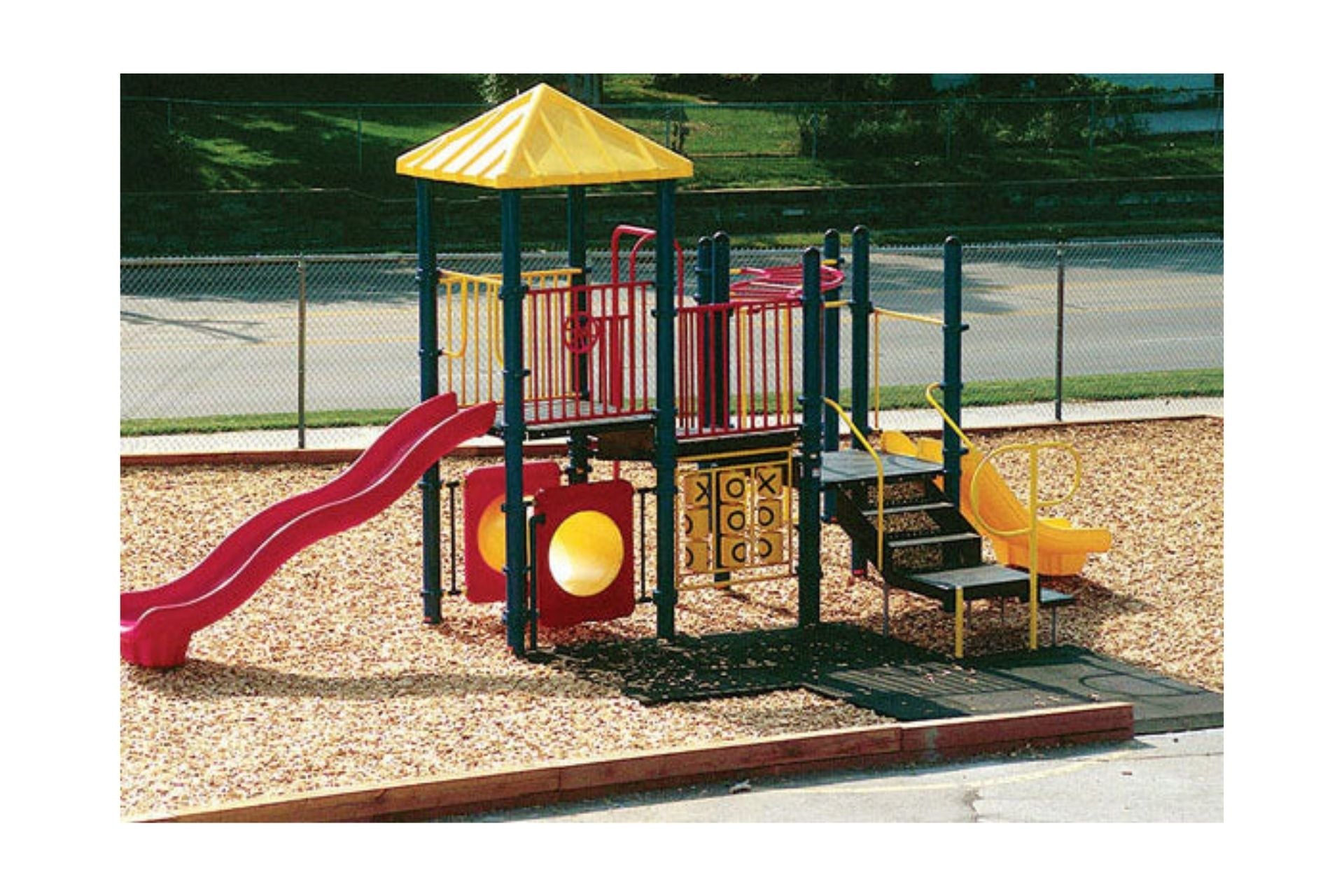 Colorful modular playground with wave and triple rail slides, tic tac toe panel, climbing elements, firepole, and yellow roof on wood chip ground.