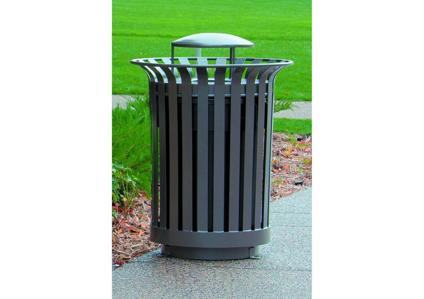 Outdoor metal trash receptacle with smooth vertical slats and rain bonnet lid on paved path near landscaping