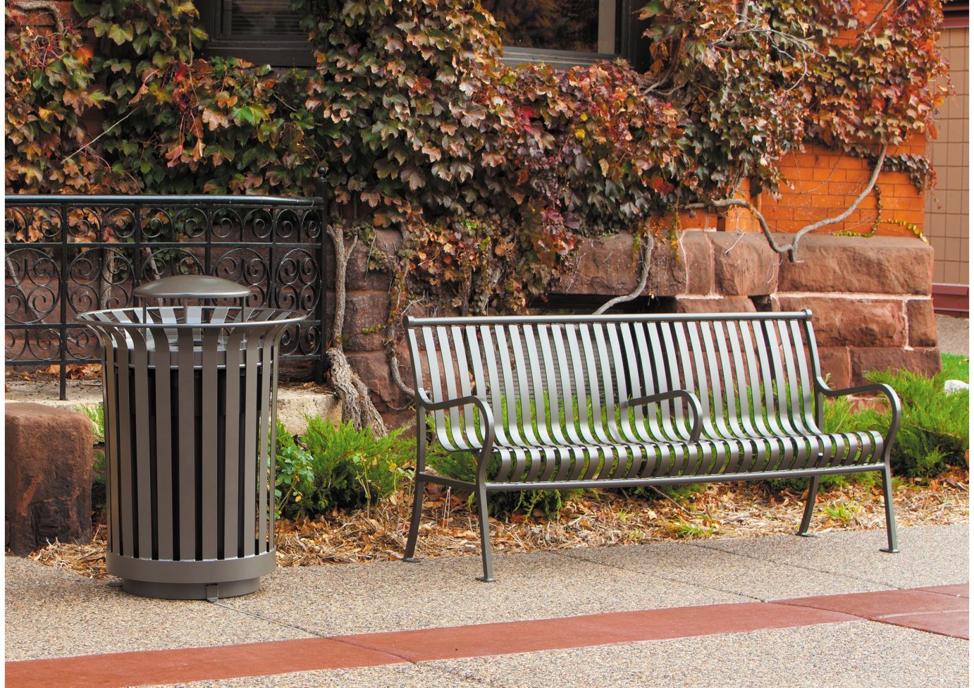 Outdoor metal trash receptacle with smooth vertical slats and rain bonnet lid beside matching metal slat bench in landscaped area