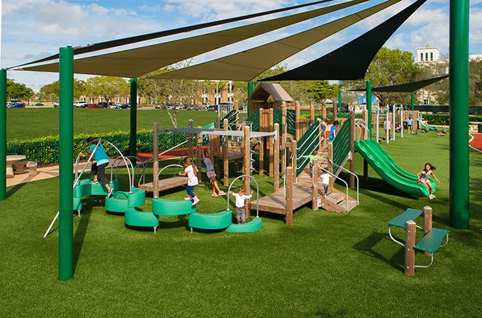 Outdoor playground with green triple wave slide, climbing net, spiral slide, shaded play structures, and children on artificial turf